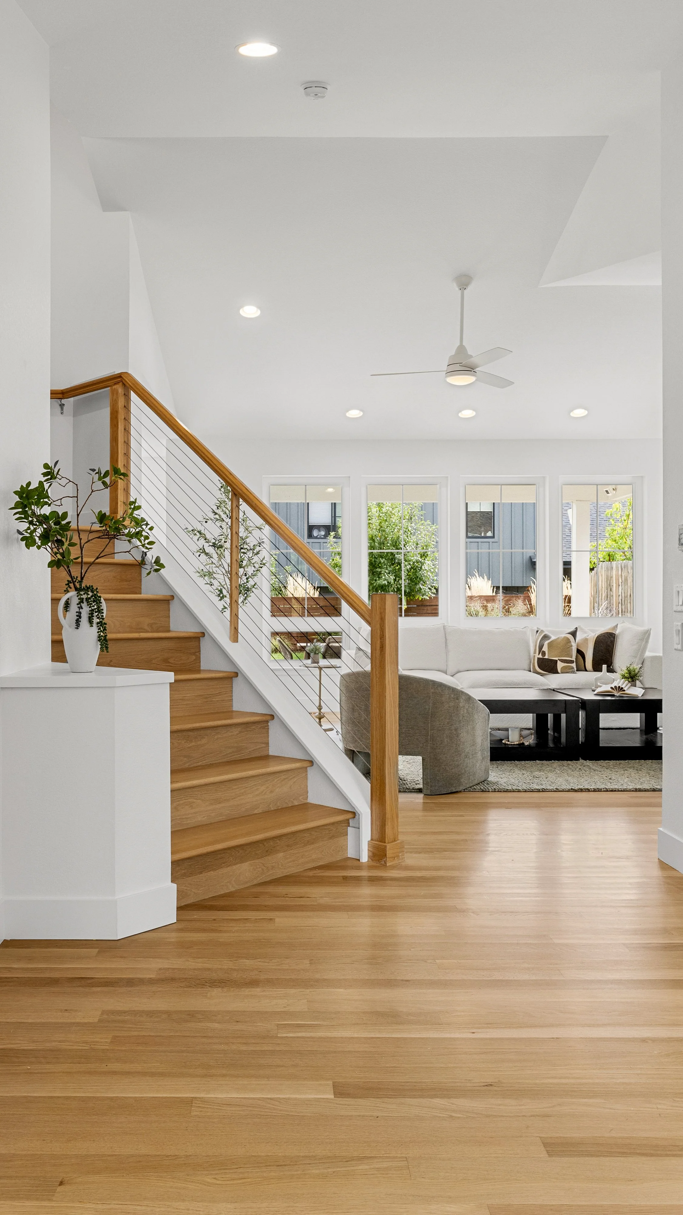 Living room with wooden staircase, white walls, ceiling fan, and large windows, with a sofa and plants.