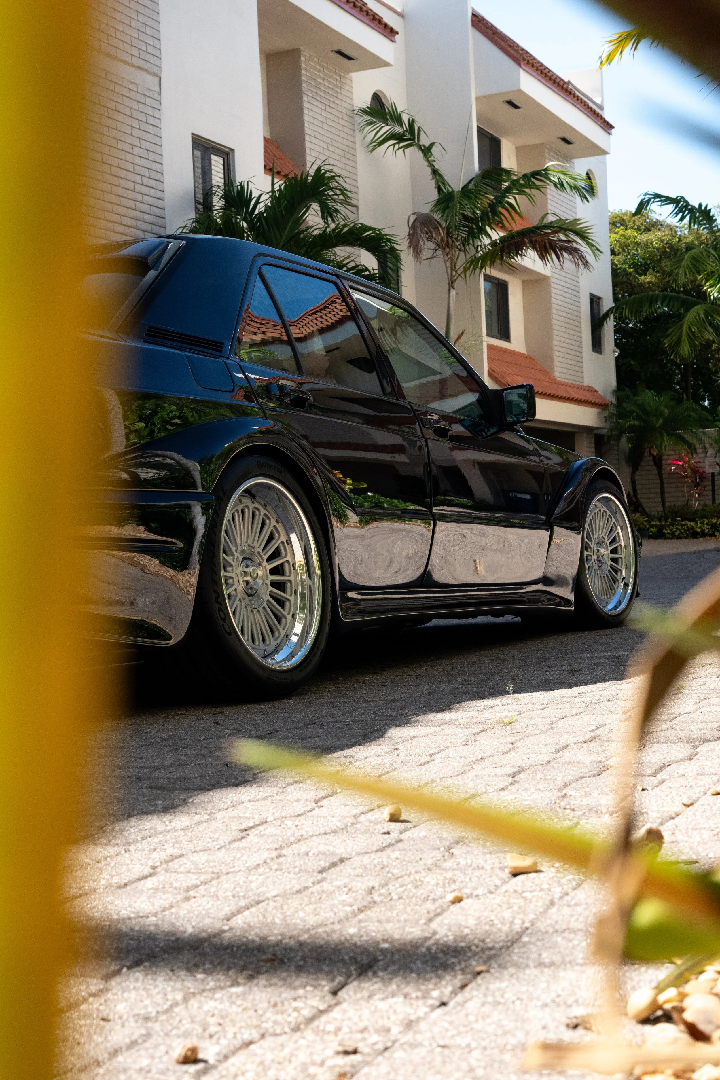 A black classic car parked in front of a modern white apartment building with palm trees, viewed through some blurred yellow and green leaves in the foreground.