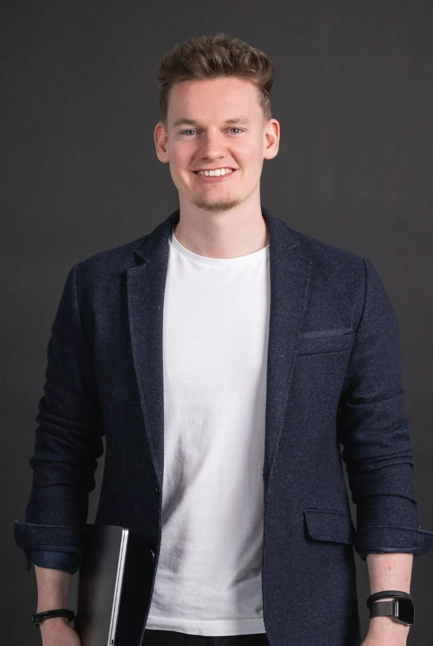 Junger Mann mit blonden Haaren und blauen Augen, trägt einen weißen T-Shirt, eine dunkelblaue Jacke und eine Armbanduhr, hält einen Laptop in der Hand, vor dunklem Hintergrund.