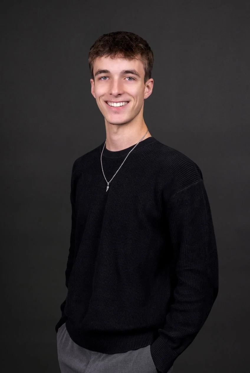 Lächelnder junger Mann trägt schwarze Sweater und graue Hosen gegen einen dunklen Hintergrund.