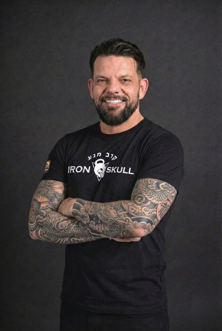Mann mit Tattoos auf den Armen, trägt schwarzes T-Shirt mit Aufdruck "Iron Skull", smilet und steht vor dunklem Hintergrund.