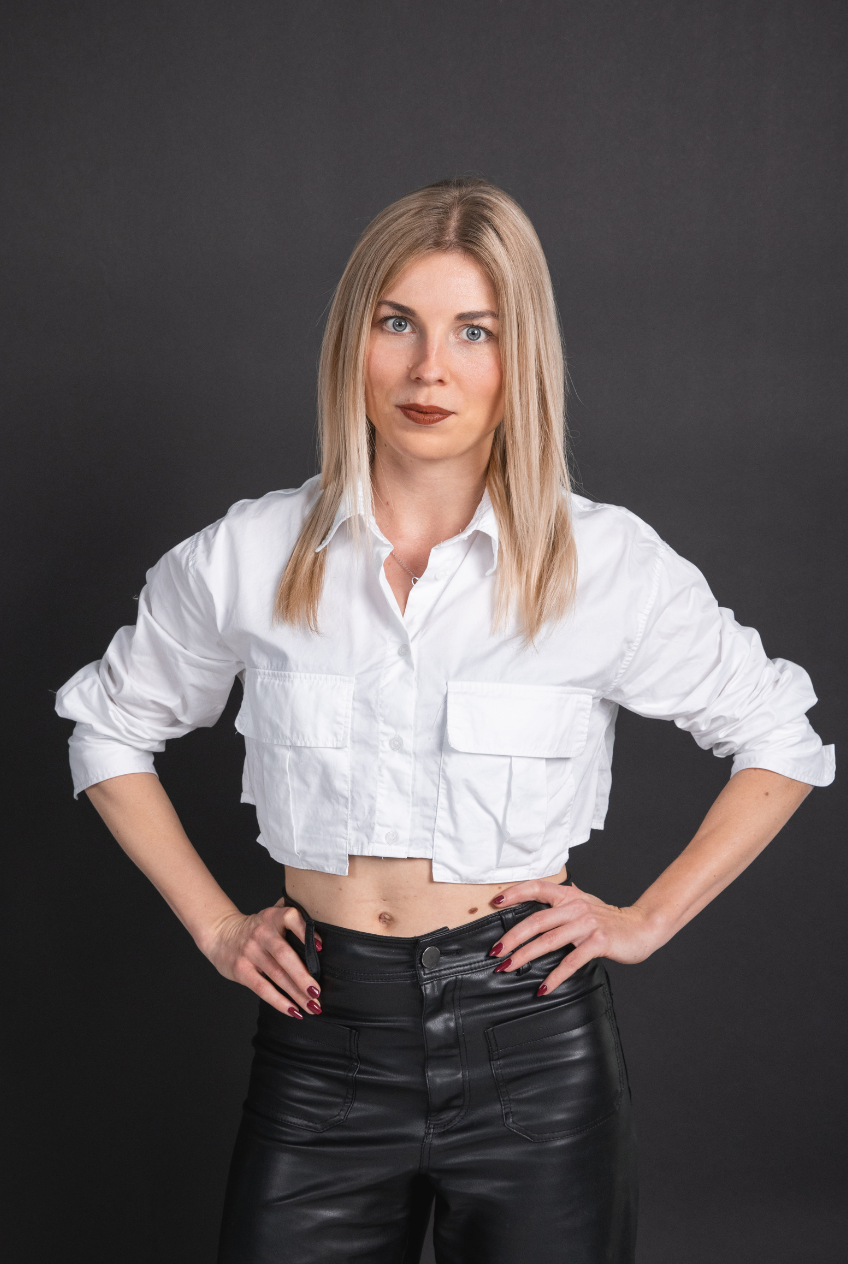 Junge Frau mit blonden Haaren, weißem Hemd und schwarzen Lederpants vor dunklem Hintergrund.