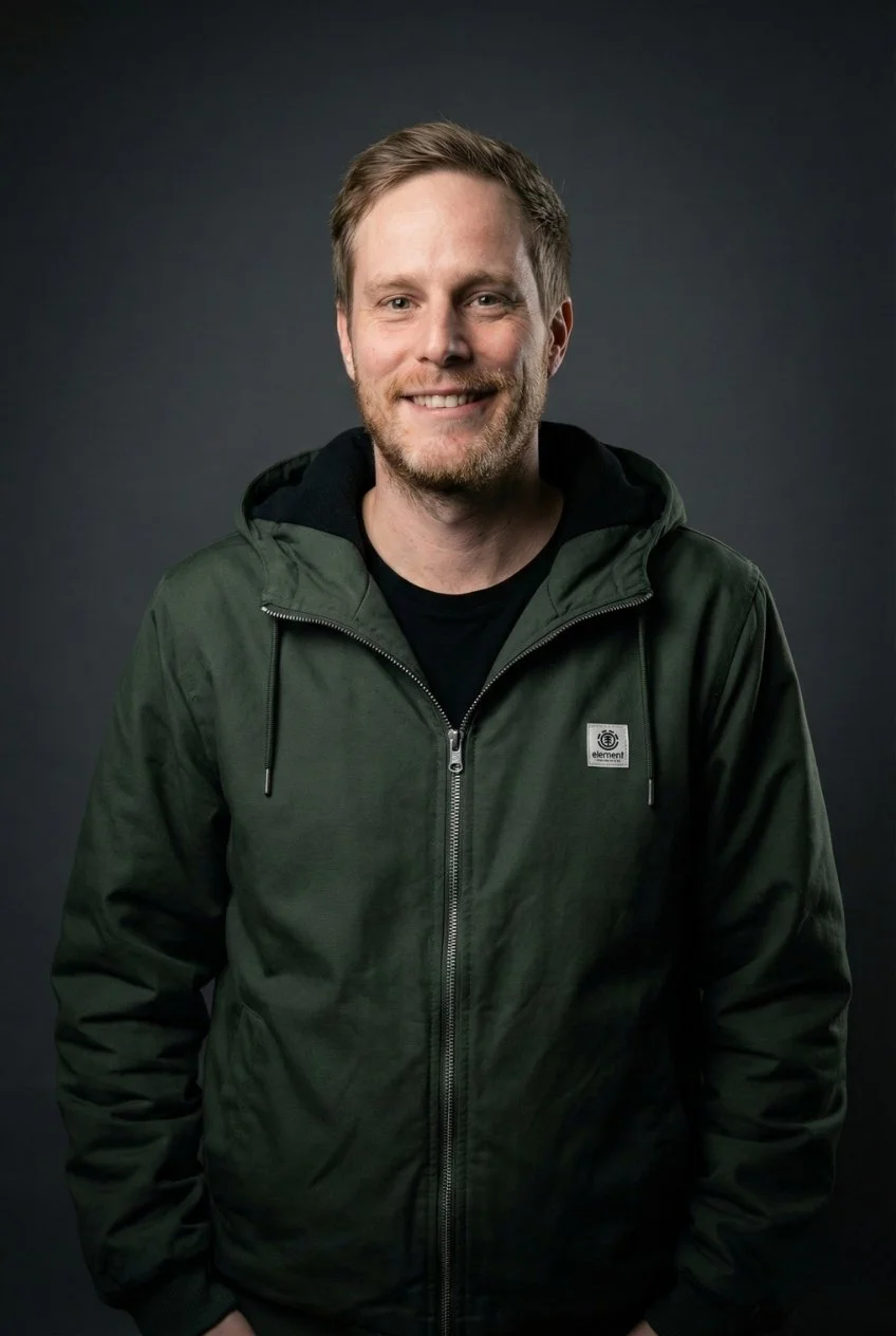 Ein Mann mit lockigem Haar und Bart, der lächelnd vor einem dunklen Hintergrund steht, trägt eine dunkle Jacke mit Reißverschluss.