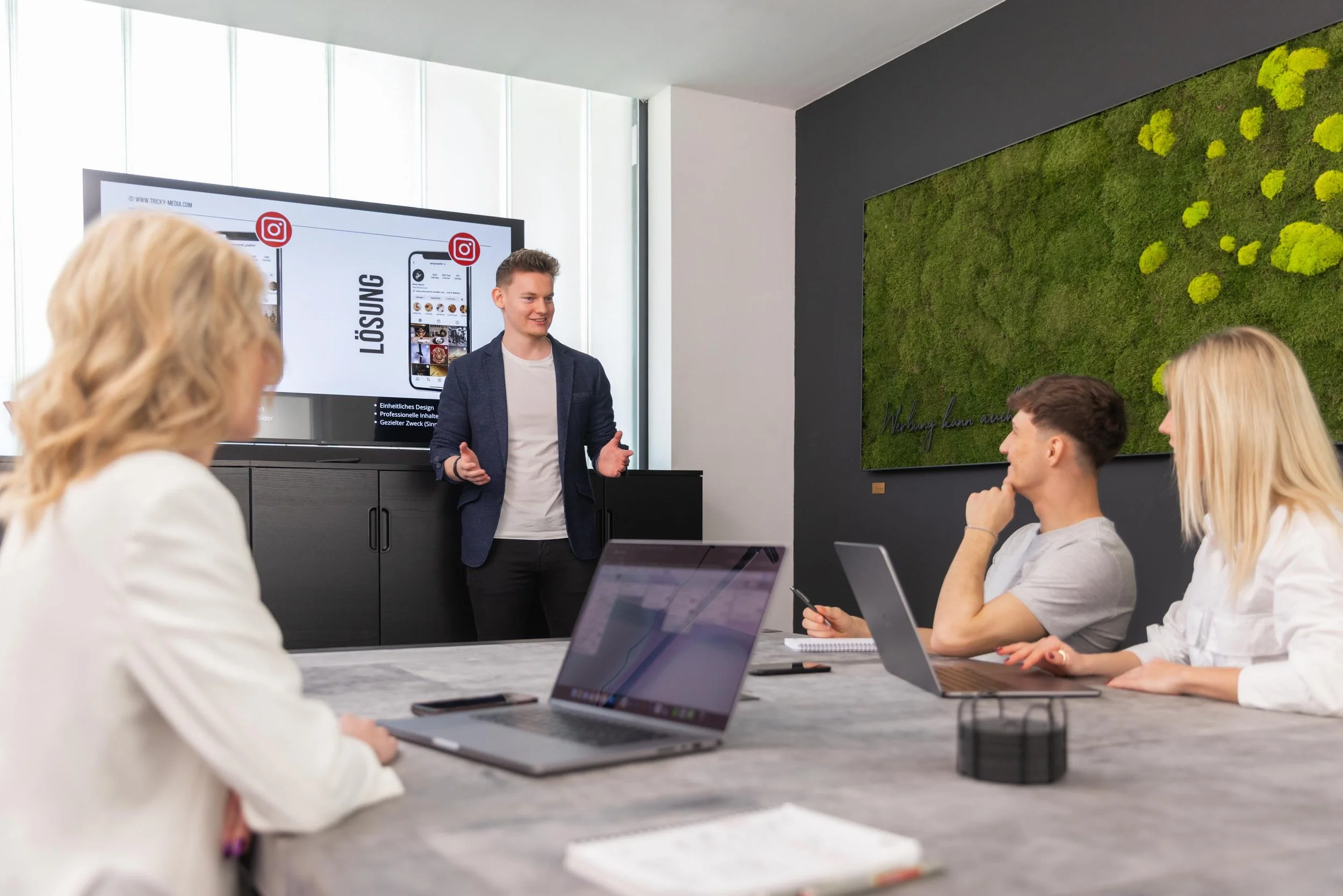 Ein Mann präsentiert eine Präsentation vor vier sitzenden Personen in einem Konferenzraum mit Laptops und Notizblock, während ein Bildschirm im Hintergrund ein Instagram-Logo und das Wort 'Lösung' zeigt.