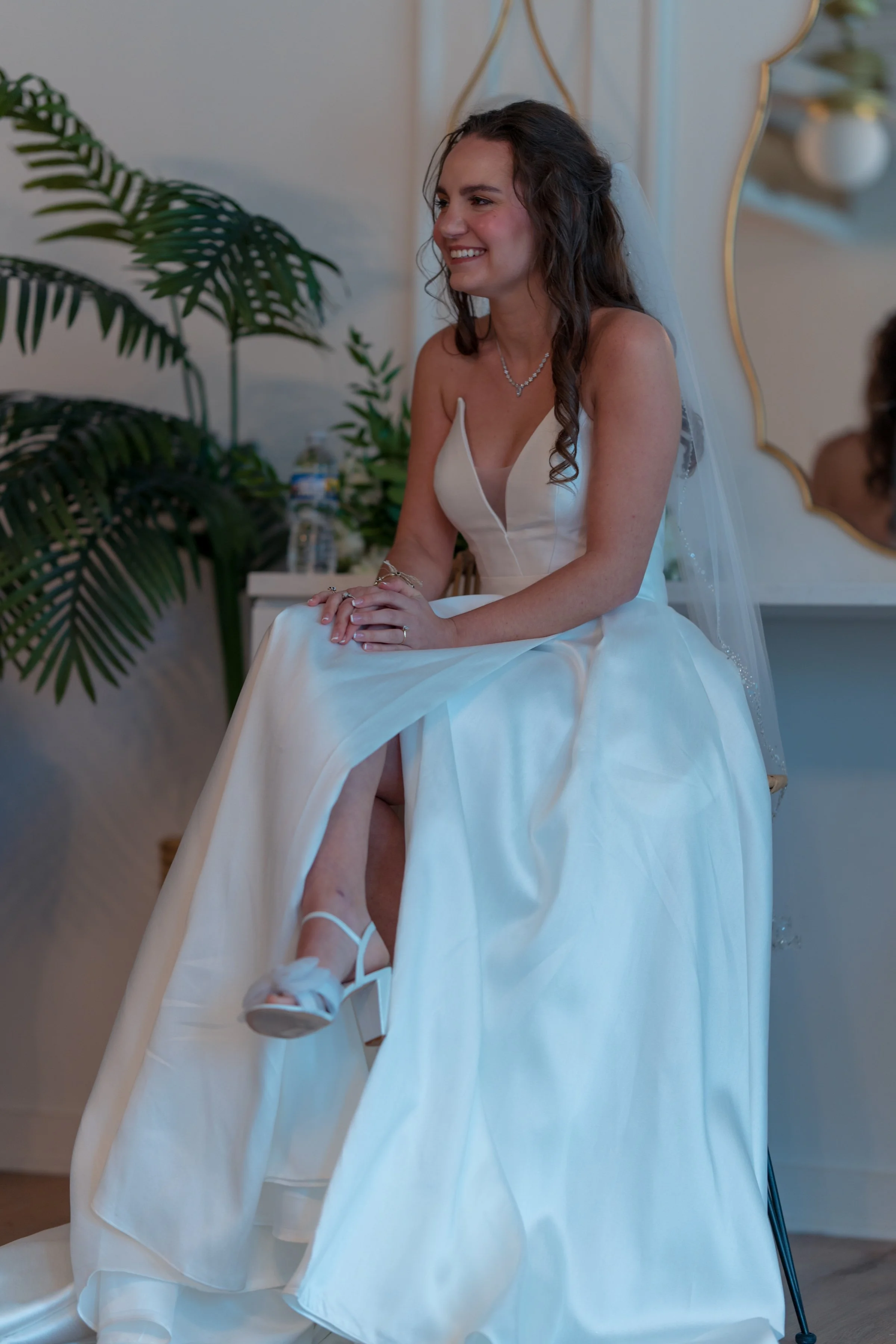 Bride sitting in a white wedding dress with a veil, smiling indoors, with plants and a mirror in the background.