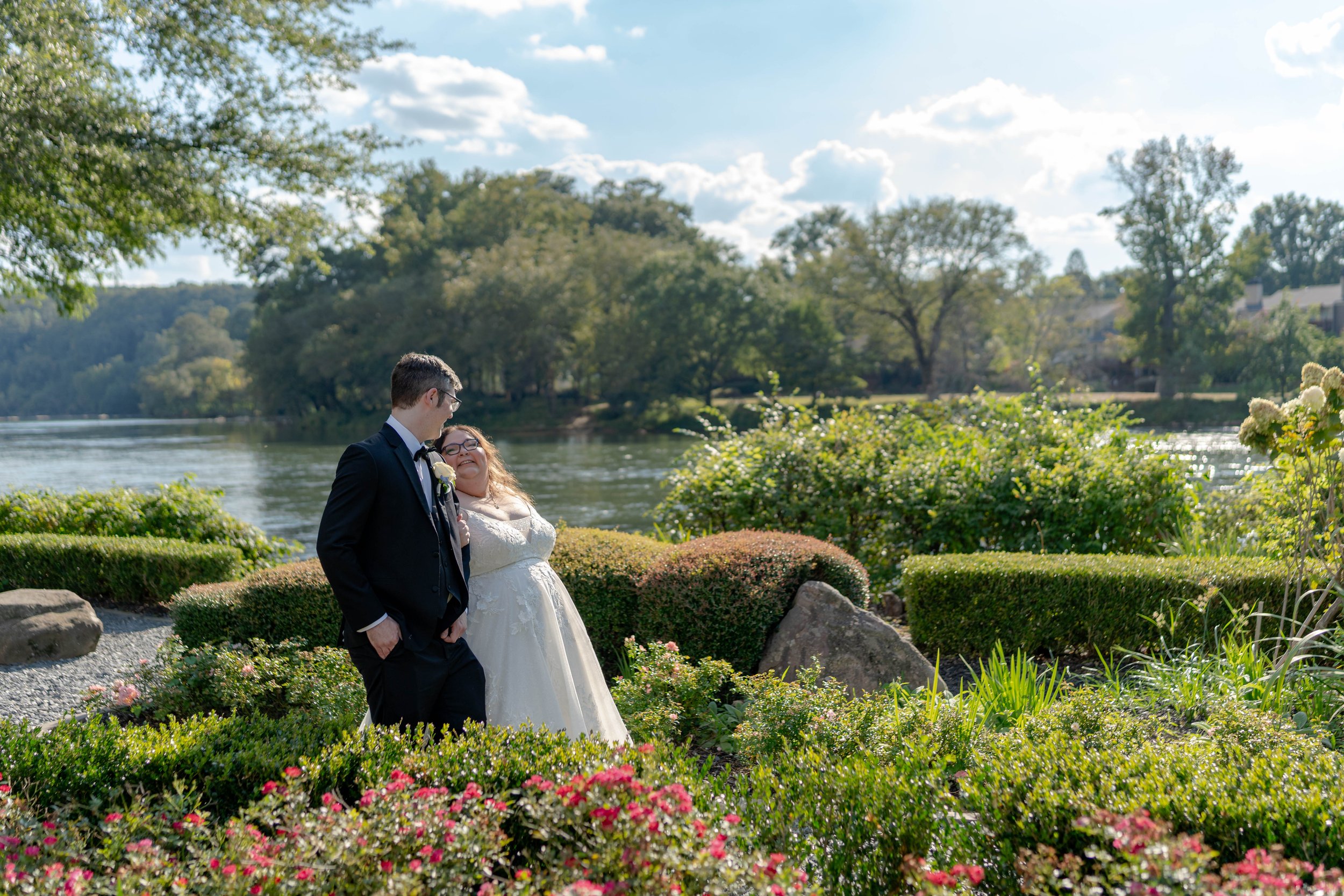 A couple dressed in wedding attire stands by a river in a park on a sunny day, surrounded by greenery and blooming flowers.