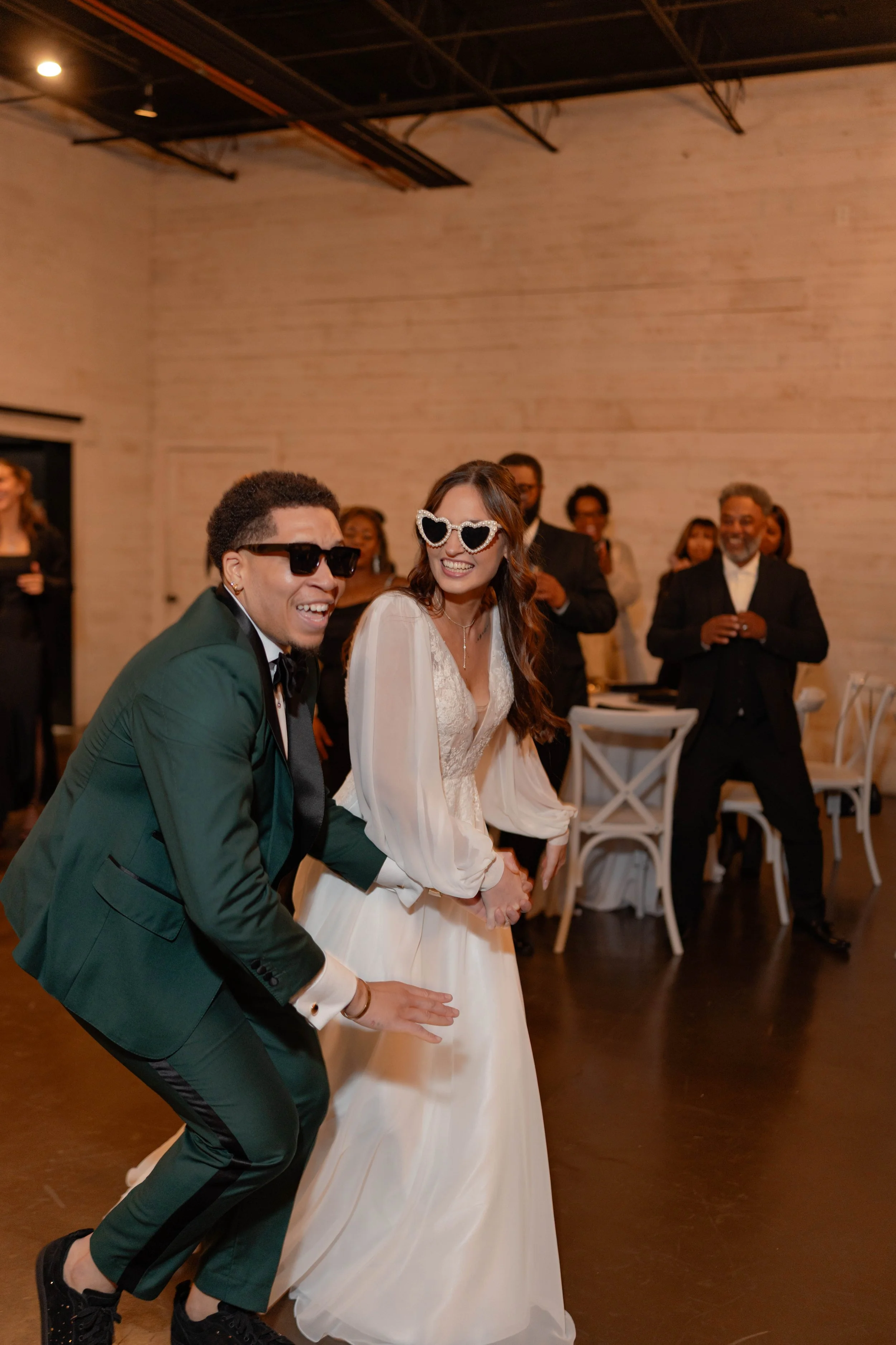 A group of people celebrating at a wedding reception. The bride and groom are in the foreground wearing sunglasses, smiling, and holding hands. The groom is dressed in a dark green tuxedo with a white shirt and black bow tie, while the bride is weari