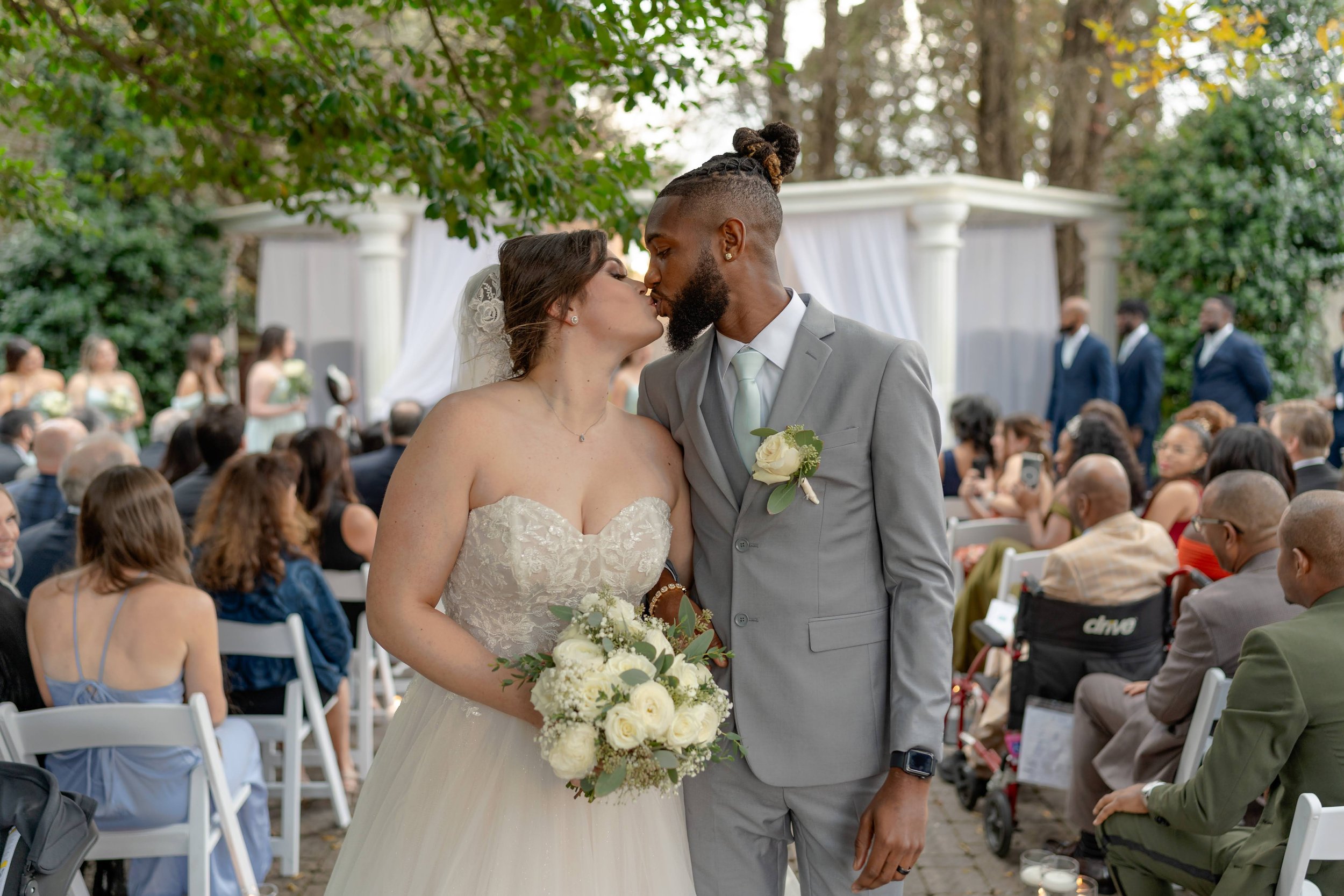 Bride and groom kissing at an outdoor wedding ceremony, with guests seated in the background.