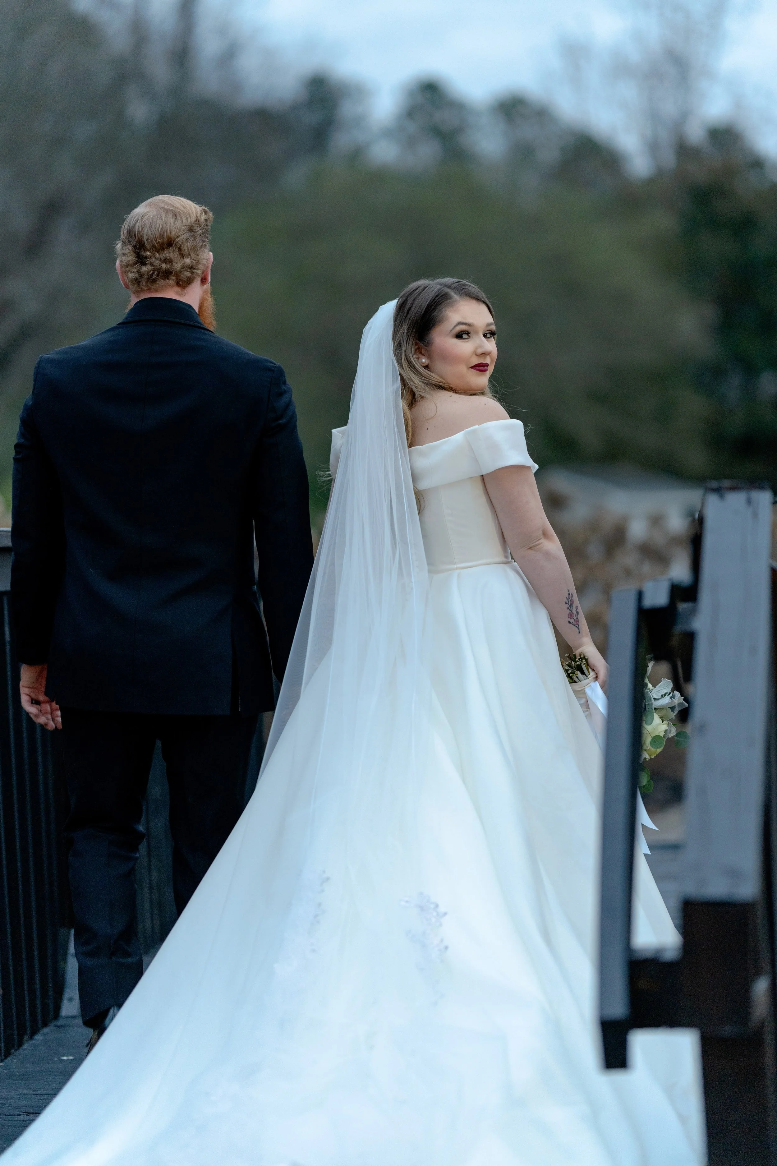 Bride in white gown with bouquet and groom in black suit walking on a wooden walkway outdoors.