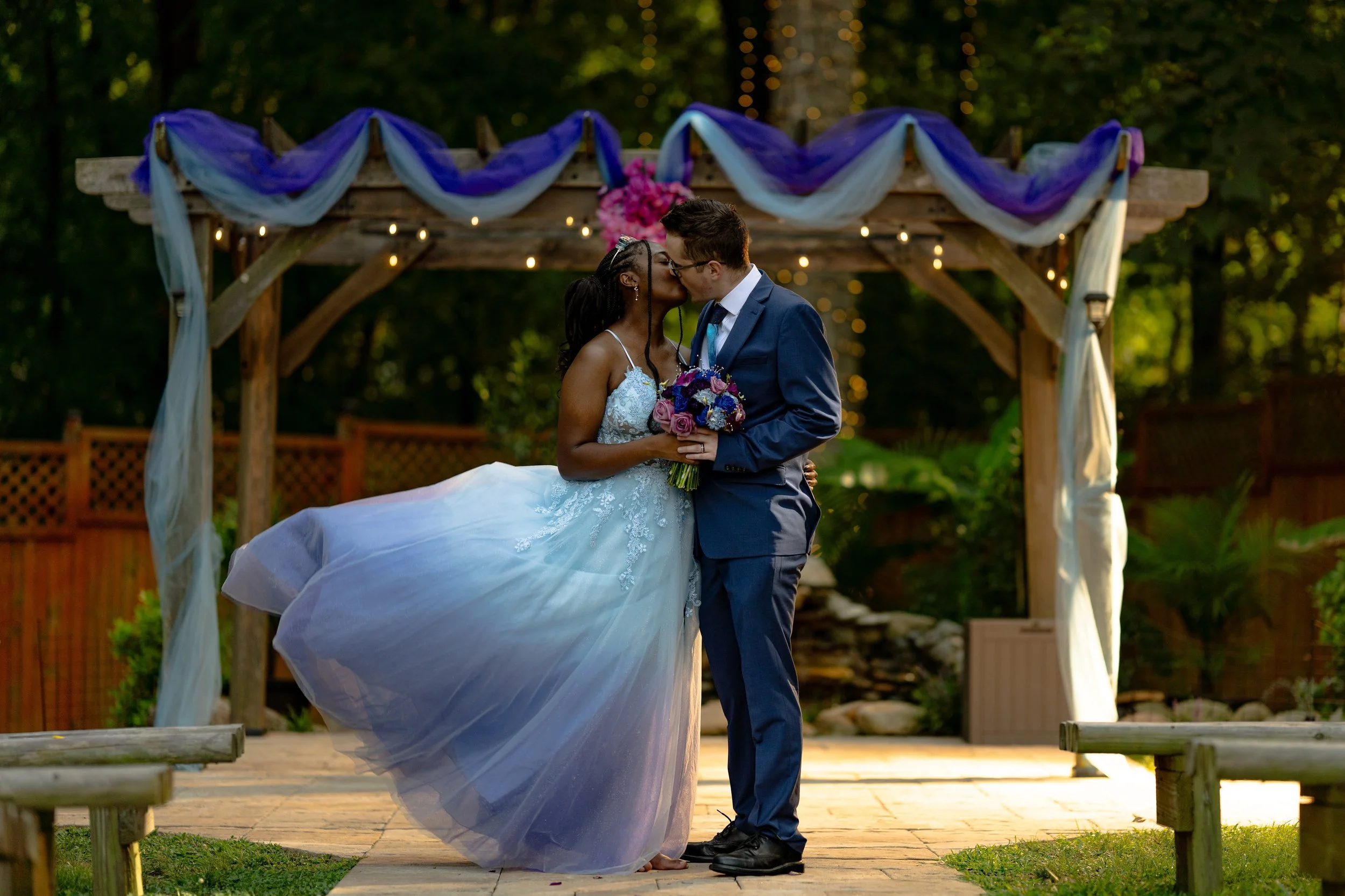 Couple kissing at outdoor wedding under decorated arch, bride in blue gown, groom in suit.