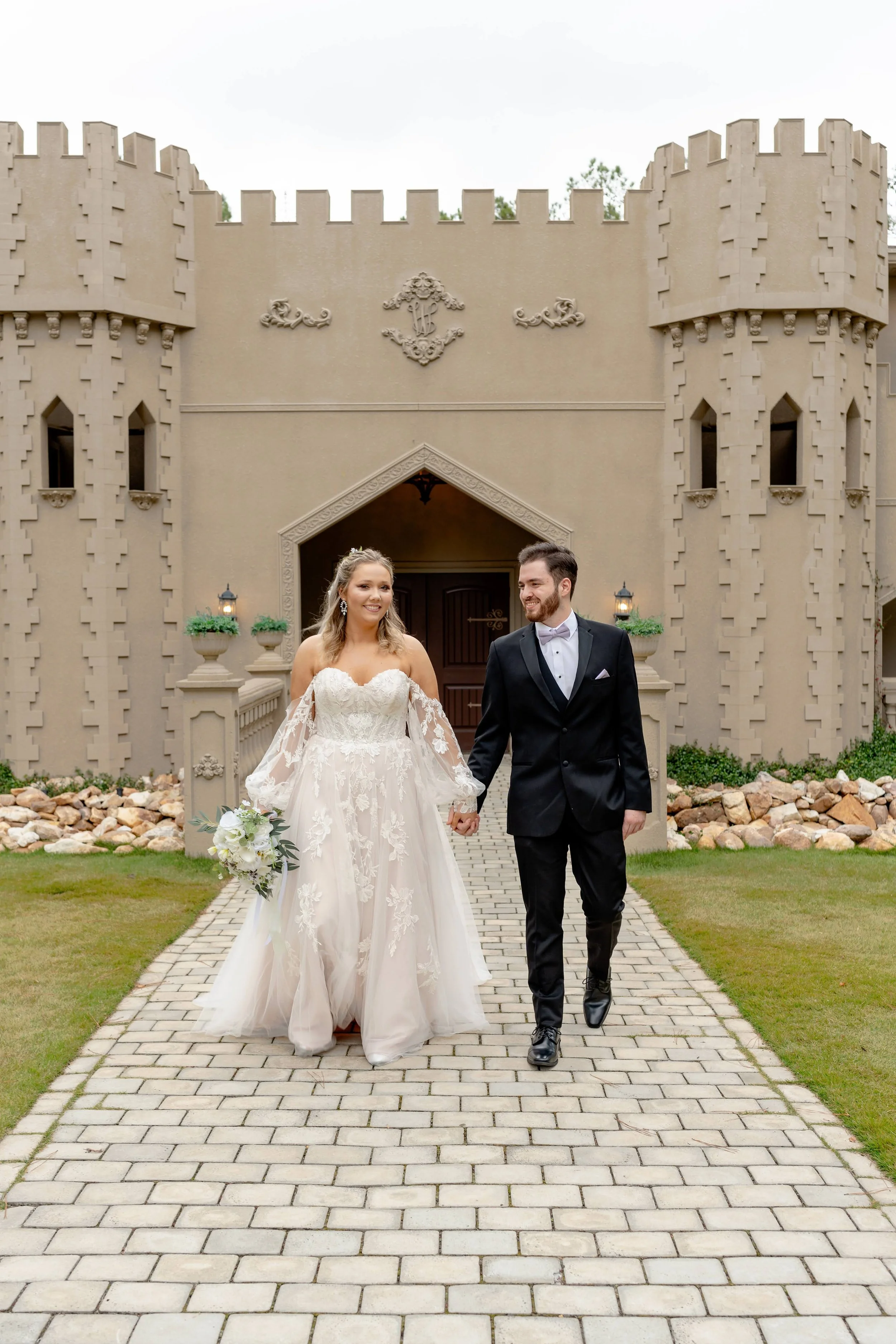 Bride and groom walking on a brick path in front of a castle-like building, with bride holding a bouquet.