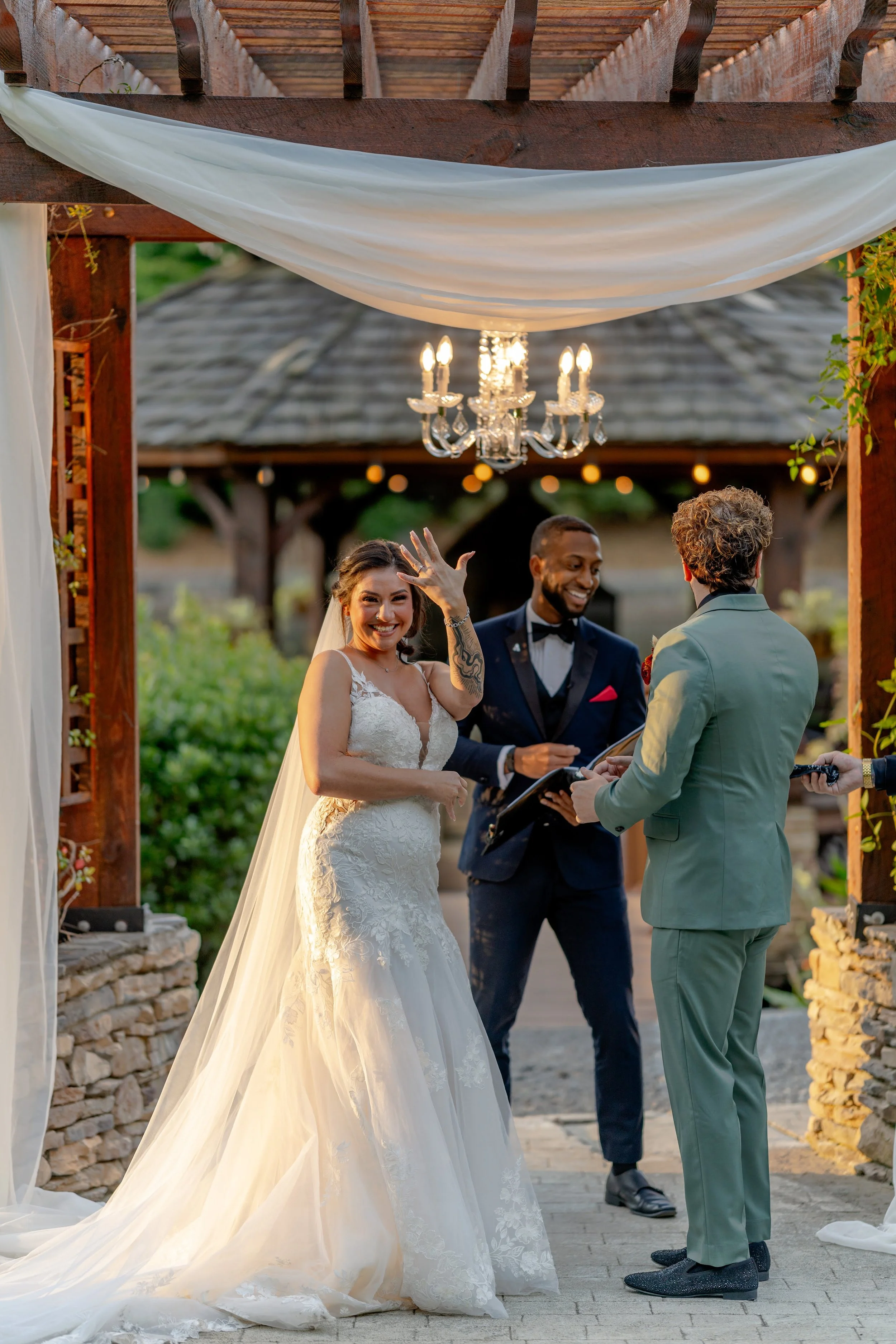 A bride and groom standing in front of an officiant under a decorated wooden arch during their wedding ceremony. The bride is wearing a white dress with a long veil and showing her wedding ring. The groom is in a green suit. A chandelier hangs above 