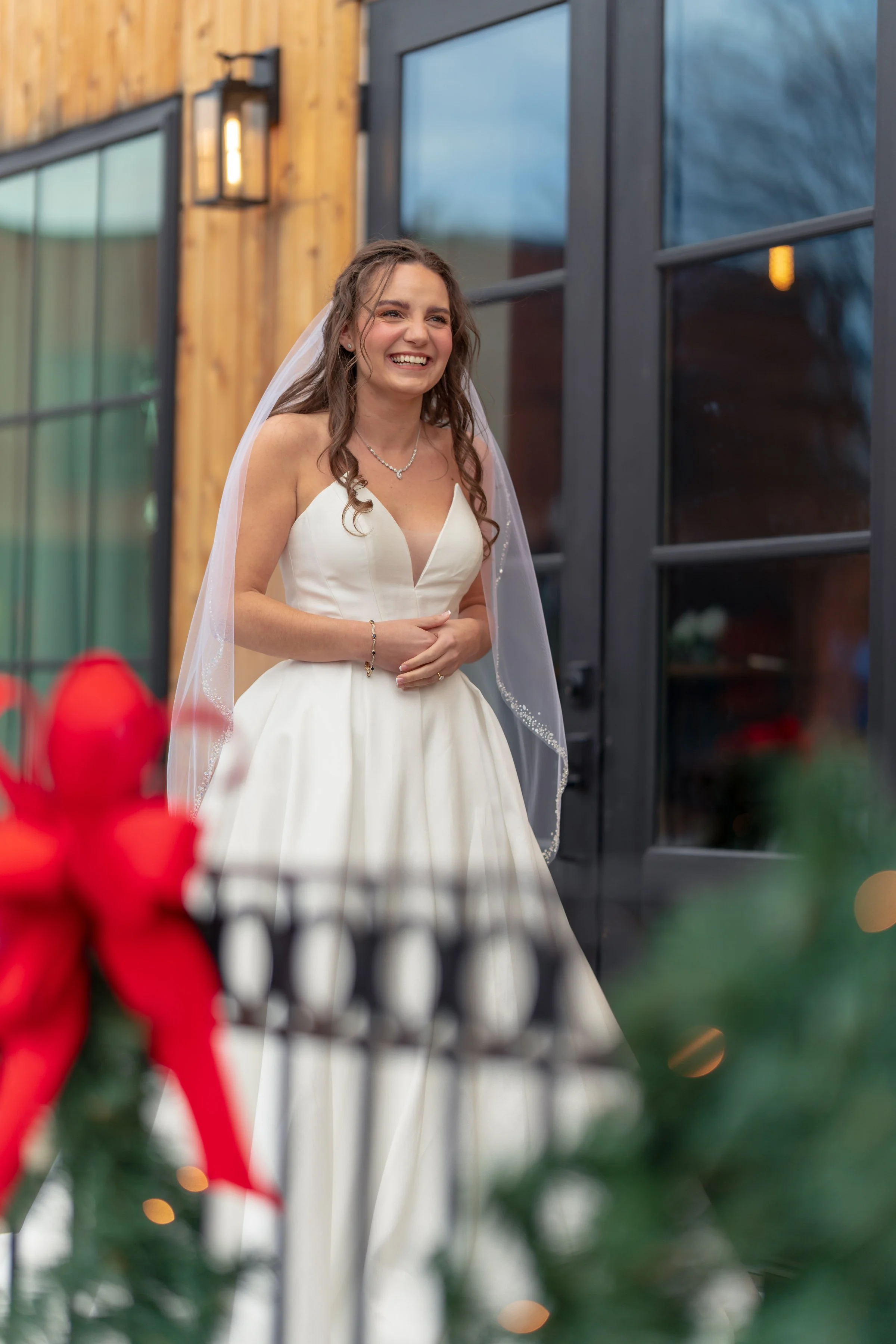 Bride smiling in white wedding dress with veil, standing outside near a decorated railing with a red bow.