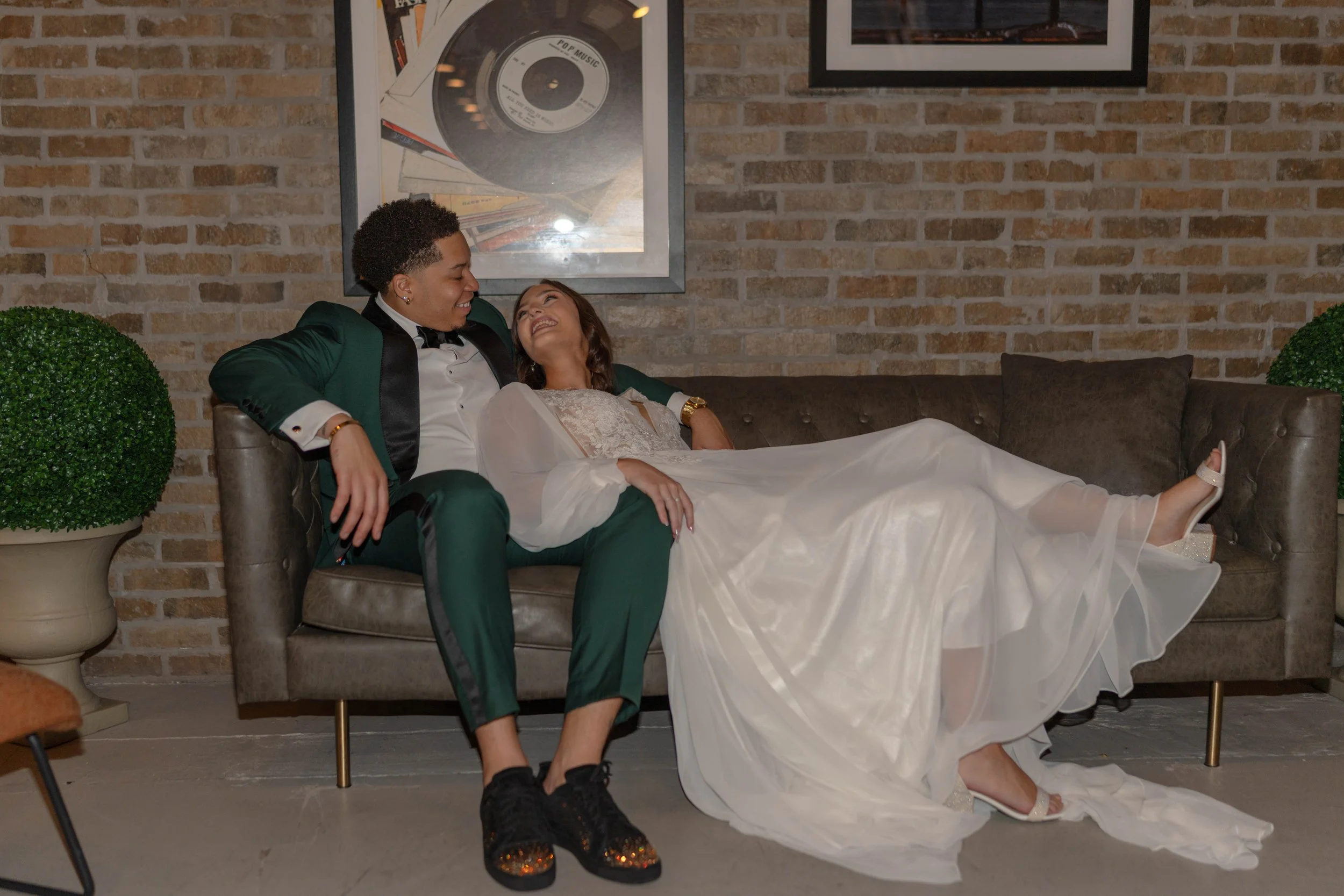A couple dressed in formal attire sitting on a vintage-style sofa with a brick wall in the background. The woman is wearing a white gown and heels, lying back with her head on the man's shoulder, both smiling and looking at each other. The man is in 