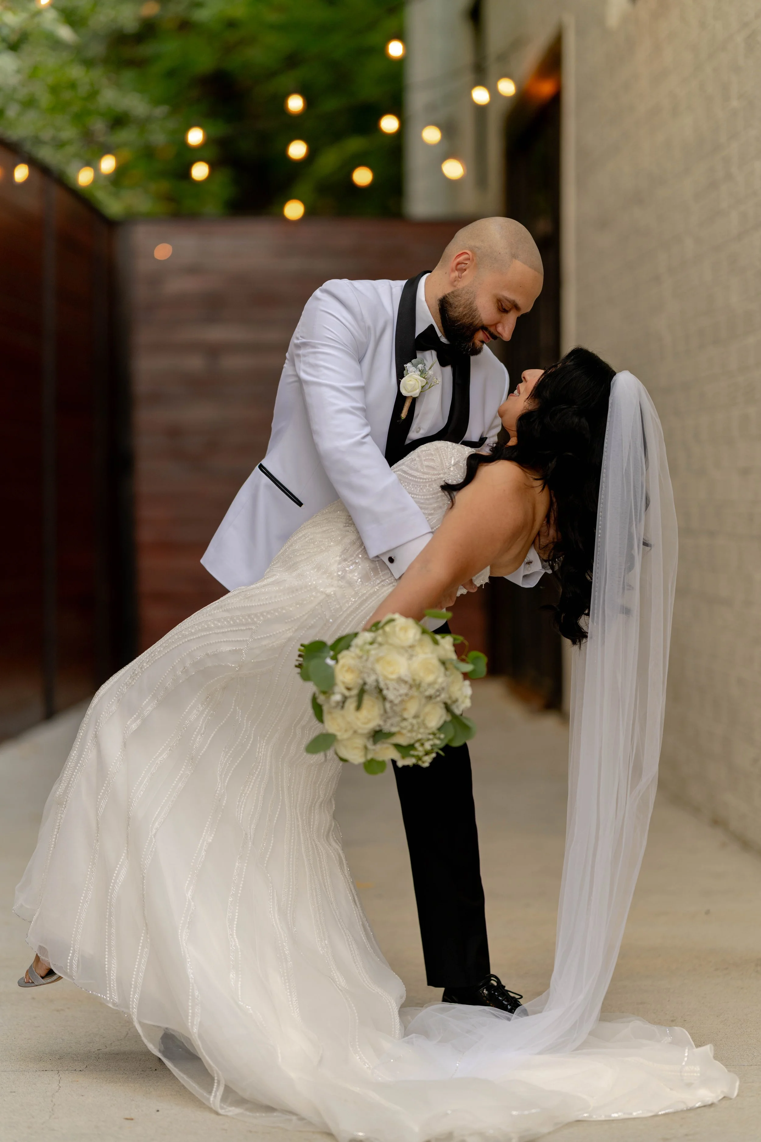 A bride in a white wedding gown with a veil and a bouquet of white roses is being dipped and kissed by a groom in a white tuxedo with a black bow tie, set against an outdoor background with string lights.