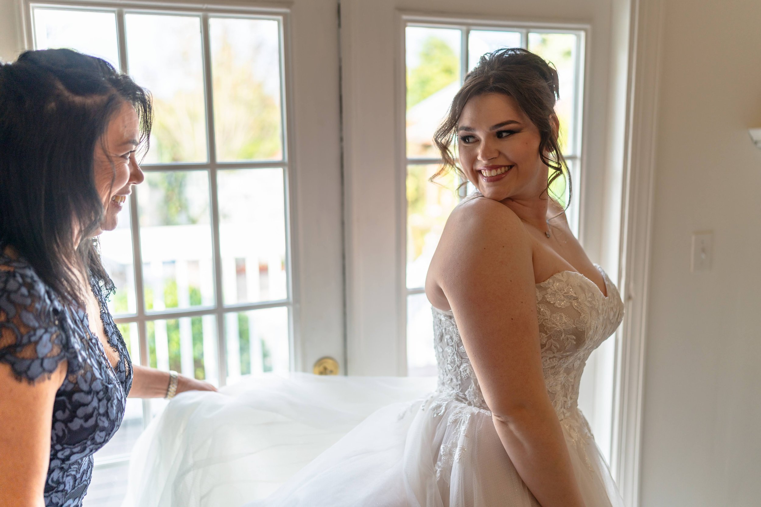 Bride in wedding dress smiling, standing near a window, with another woman helping with the gown.