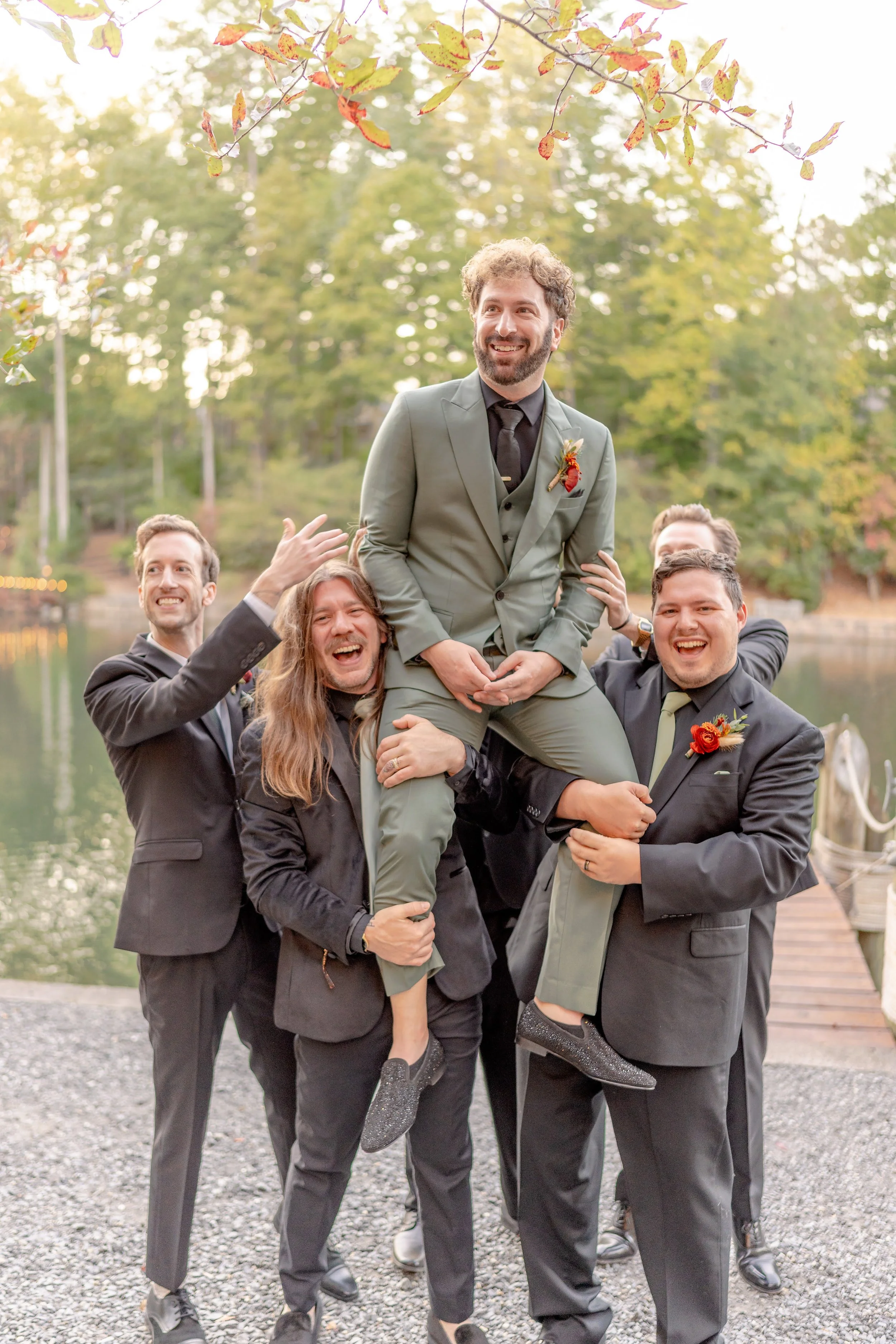 Groomsmen lifting the groom in outdoor wedding setting, dressed in suits.