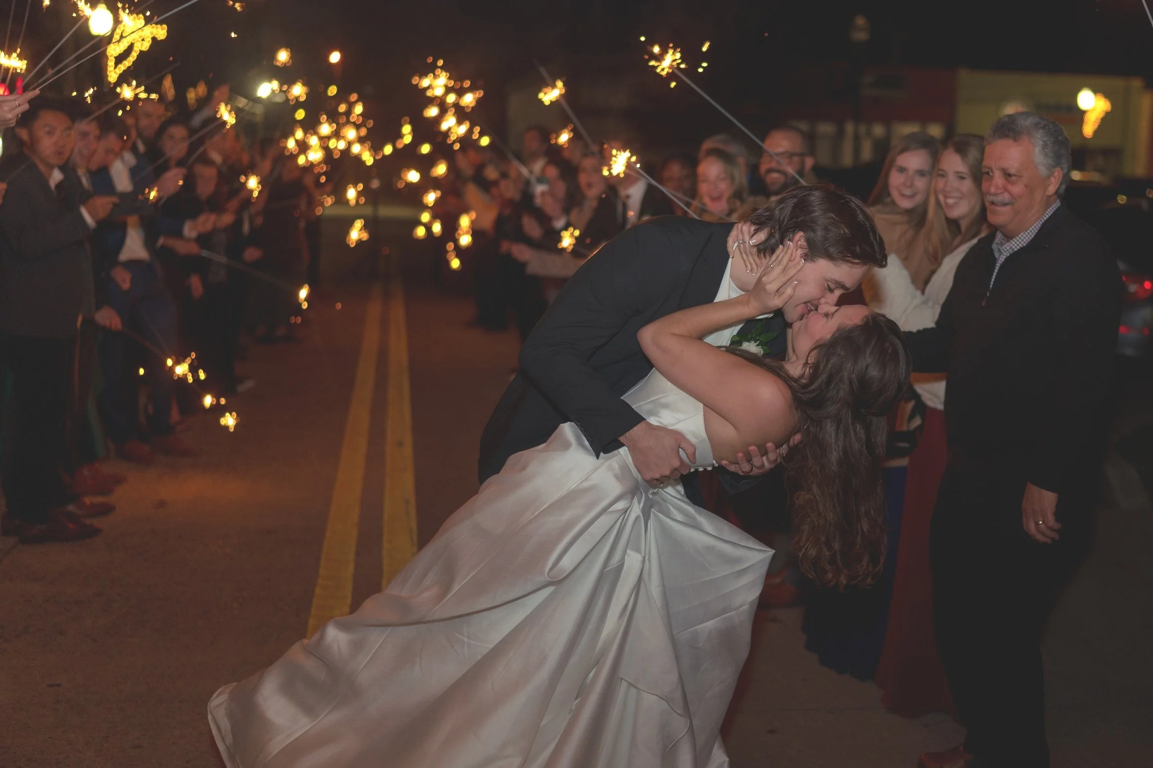 Bride and groom kissing during sparkler send-off at night, surrounded by guests holding sparklers.