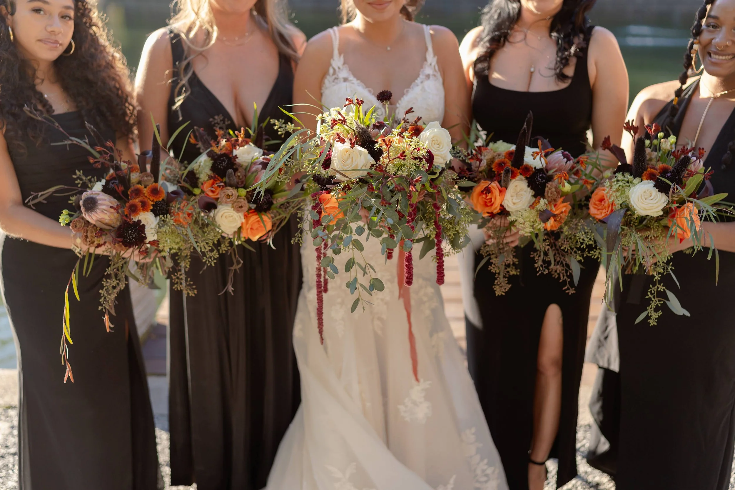 Bridal party holding colorful flower bouquets with white, orange, red flowers and greenery, bridesmaids in black dresses, bride in white lace dress.