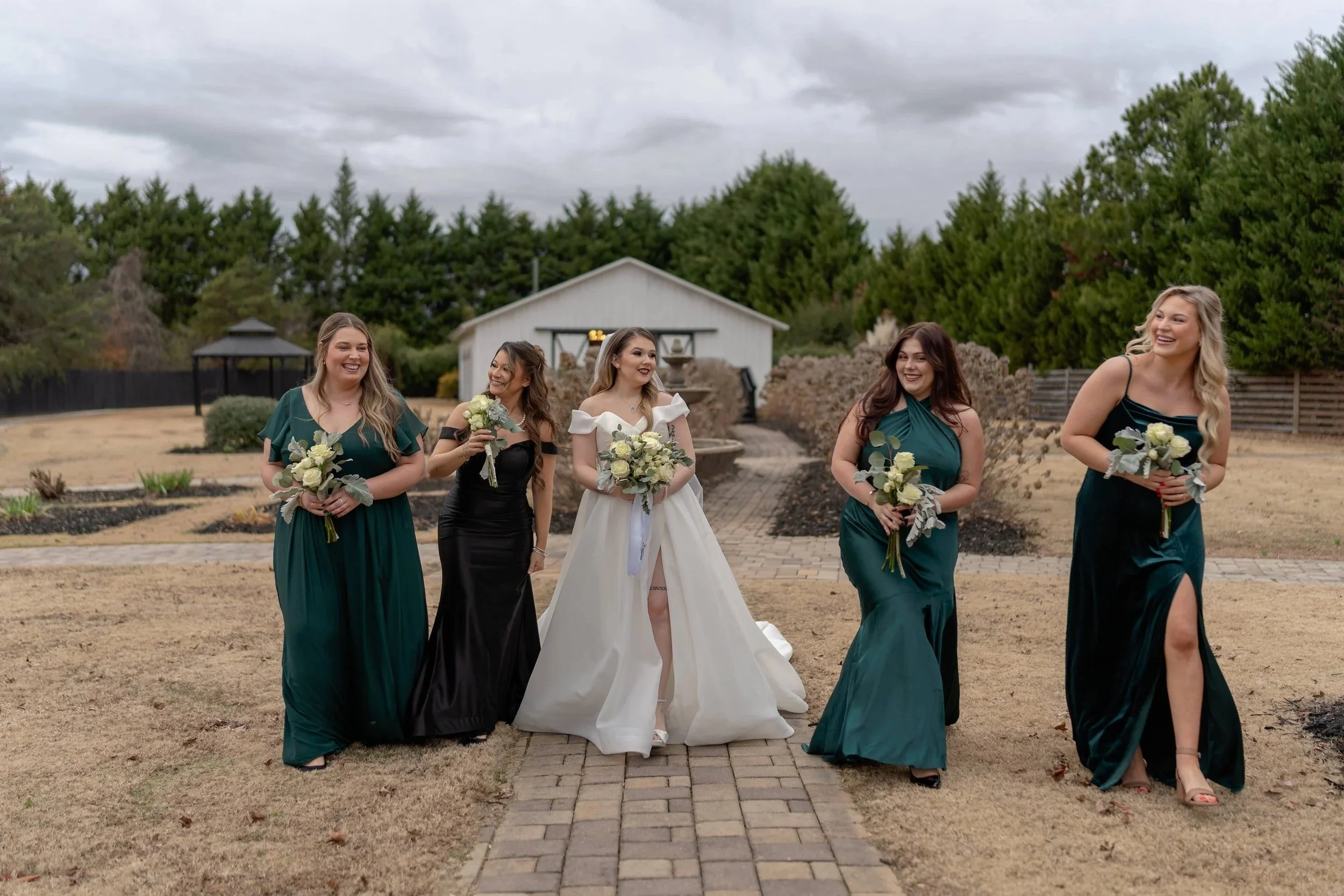 A bride and four bridesmaids walking outdoors on a cloudy day, each holding a bouquet of white flowers with greenery. The bride is wearing a white wedding gown with off-shoulder sleeves and the bridesmaids are wearing dark green and black dresses. They are smiling and appear happy.