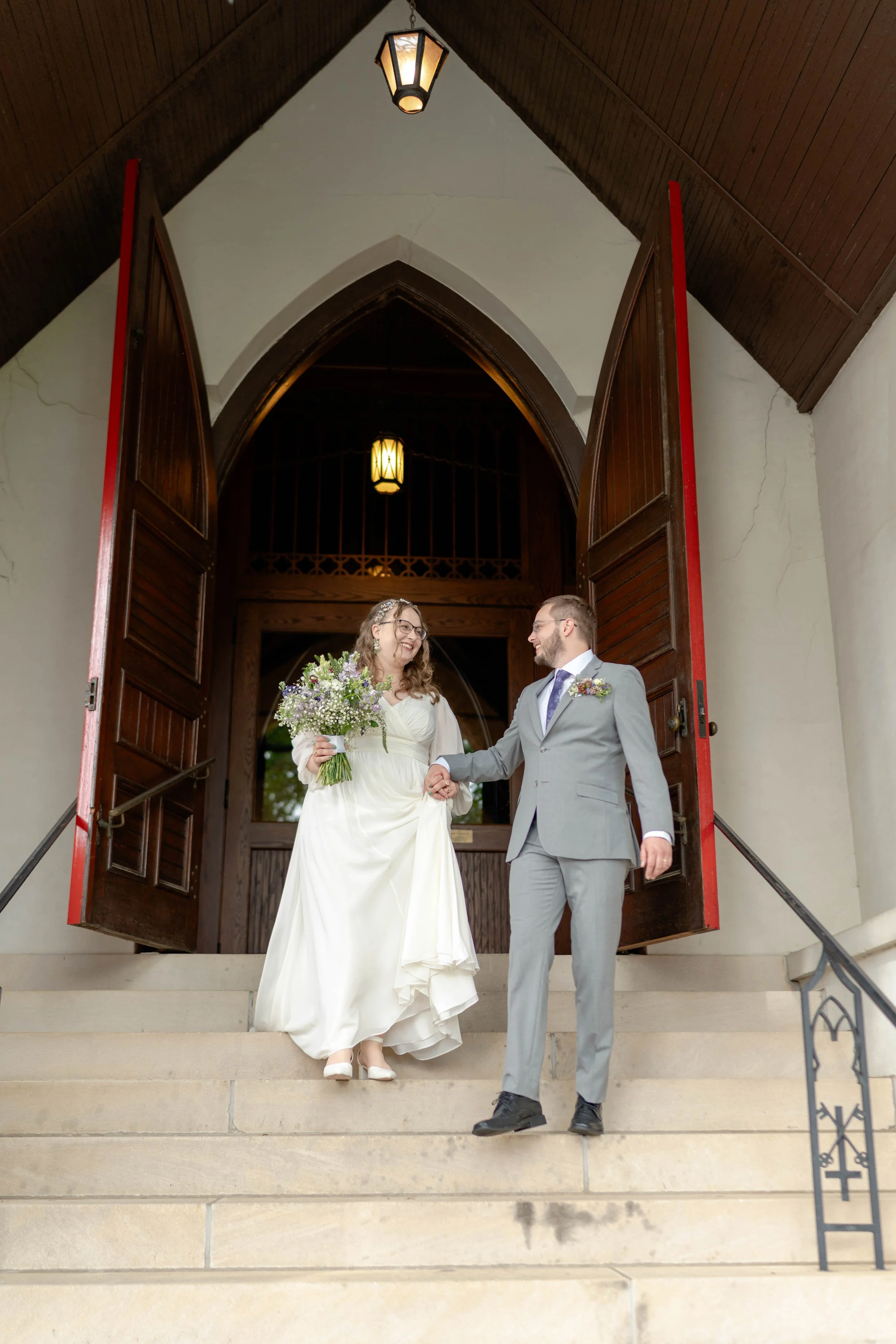A bride and groom are standing on the steps of a church or chapel, holding hands and smiling at each other. The bride is wearing a white dress and holding a large bouquet of flowers, while the groom is dressed in a light gray suit with a boutonniere.
