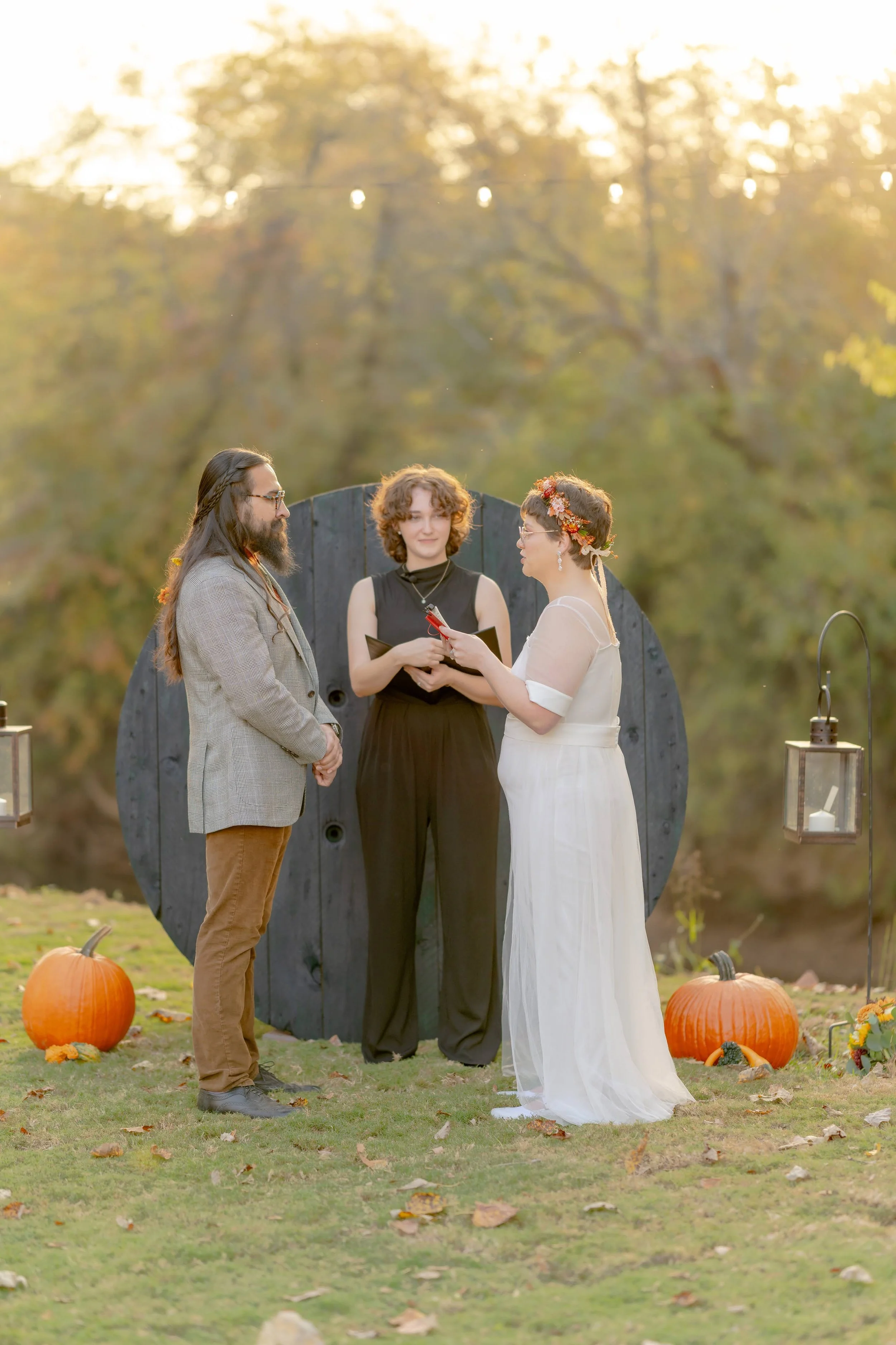 Outdoor wedding ceremony with a couple exchanging vows, officiated by a person in black. Decor includes pumpkins and lanterns, with autumn foliage in the background.