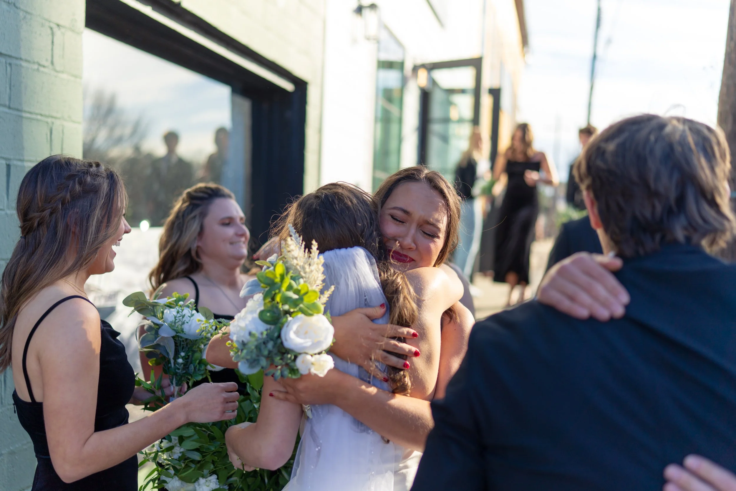 A group of people celebrating outside, with a woman in a wedding dress hugging another woman, who appears emotional. Others around them are smiling, and two of them hold bouquets. The setting is sunny, near a building with large windows.