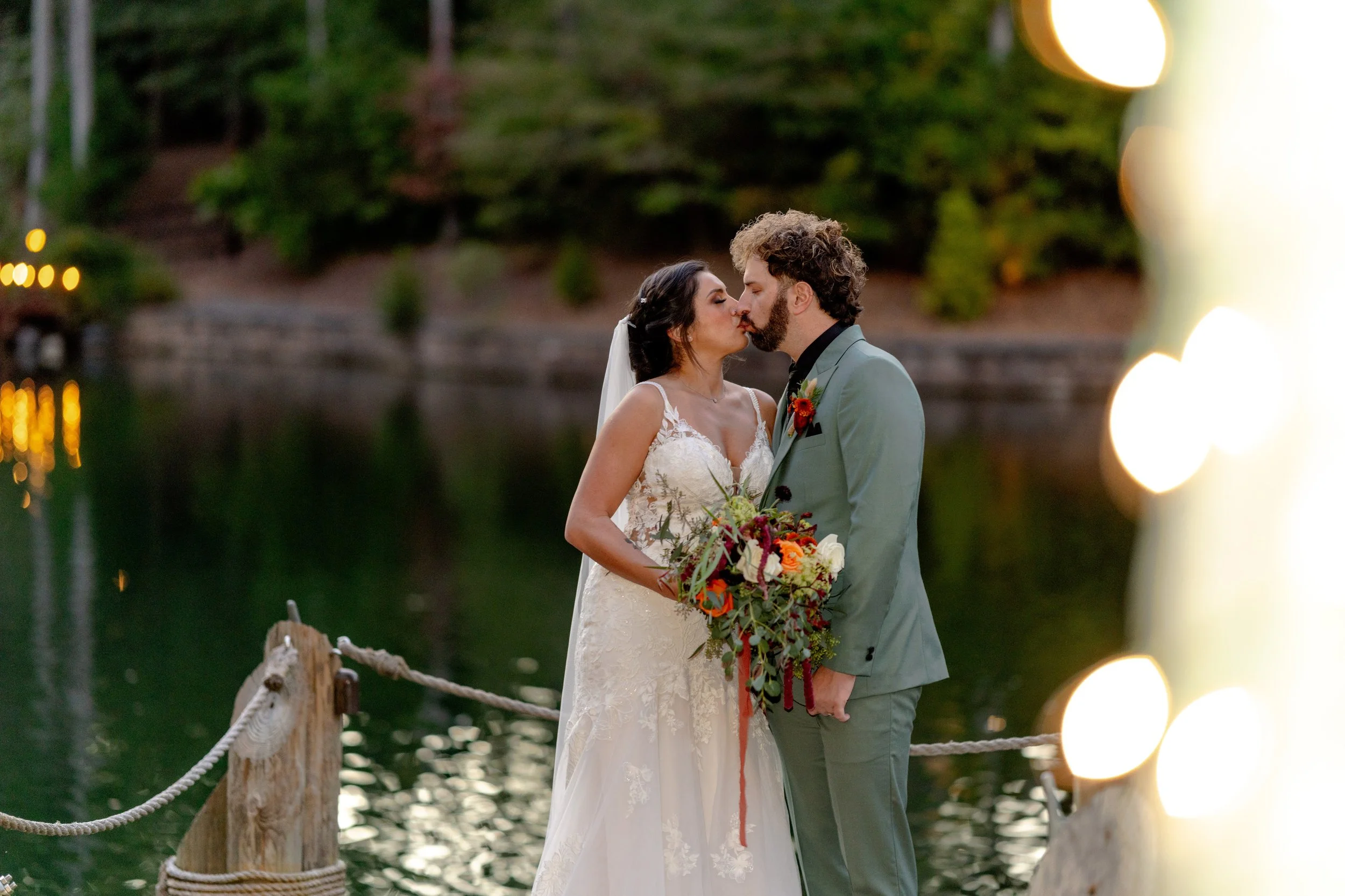 Bride and groom kissing near a lake, bride in a white wedding dress holding a bouquet, groom in a light-colored suit with a boutonniere, surrounded by greenery and soft lighting.
