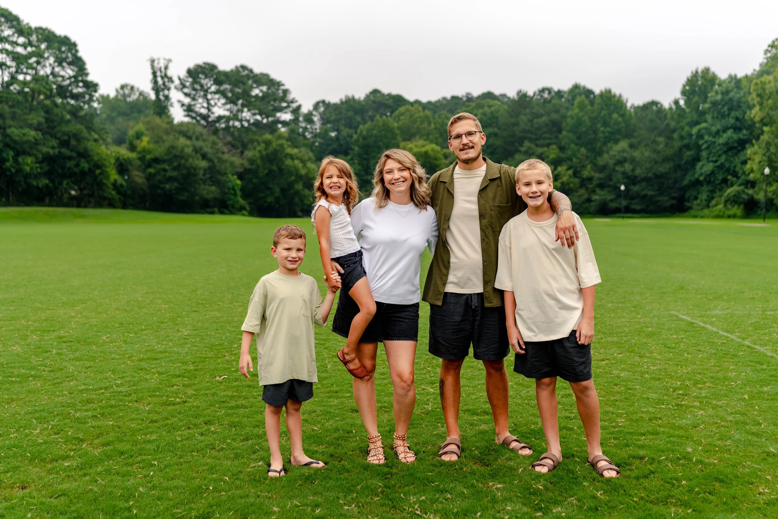A family of five standing on a green field with trees in the background, smiling at the camera.