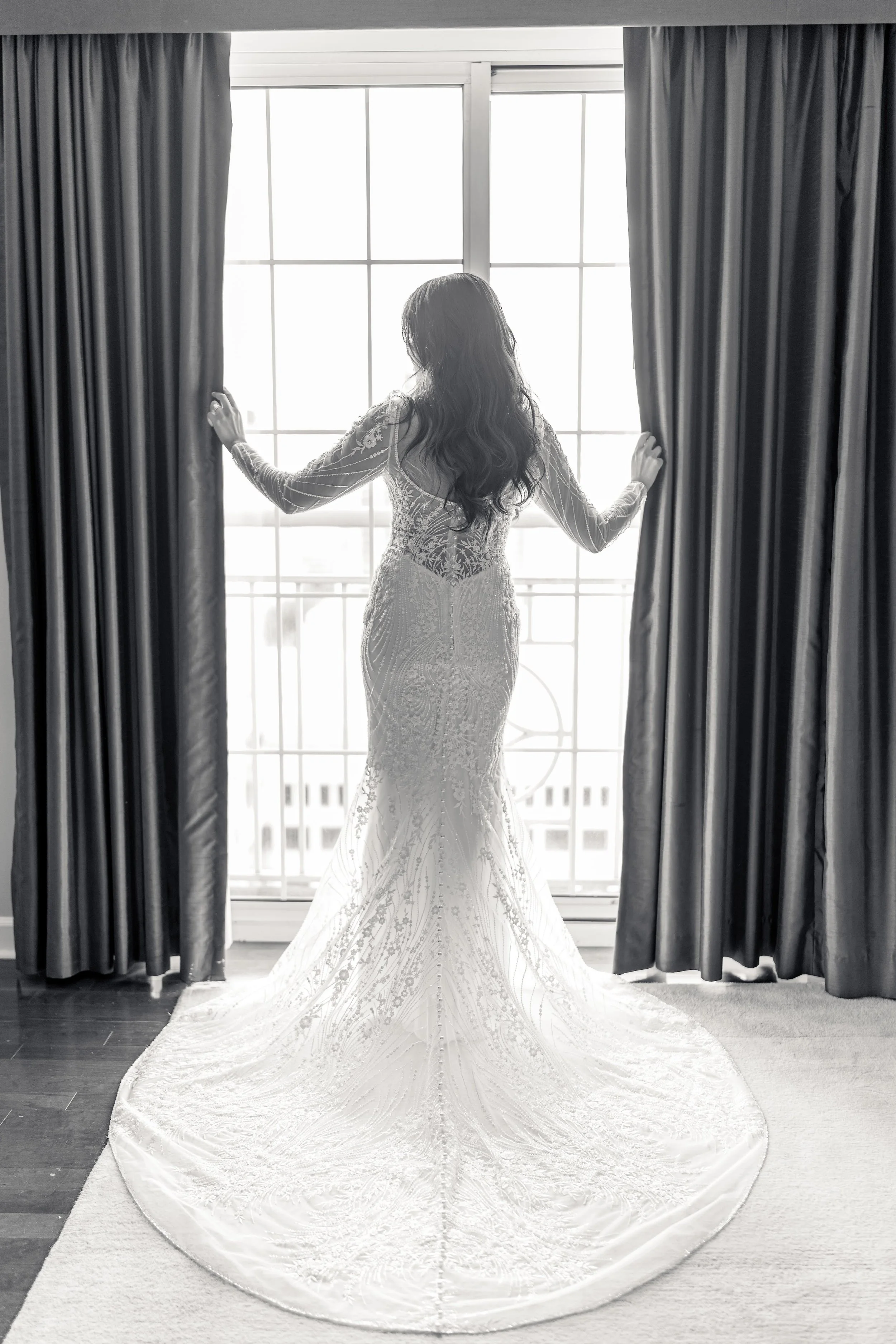 Bride in lace wedding dress standing at window with curtains, back view.