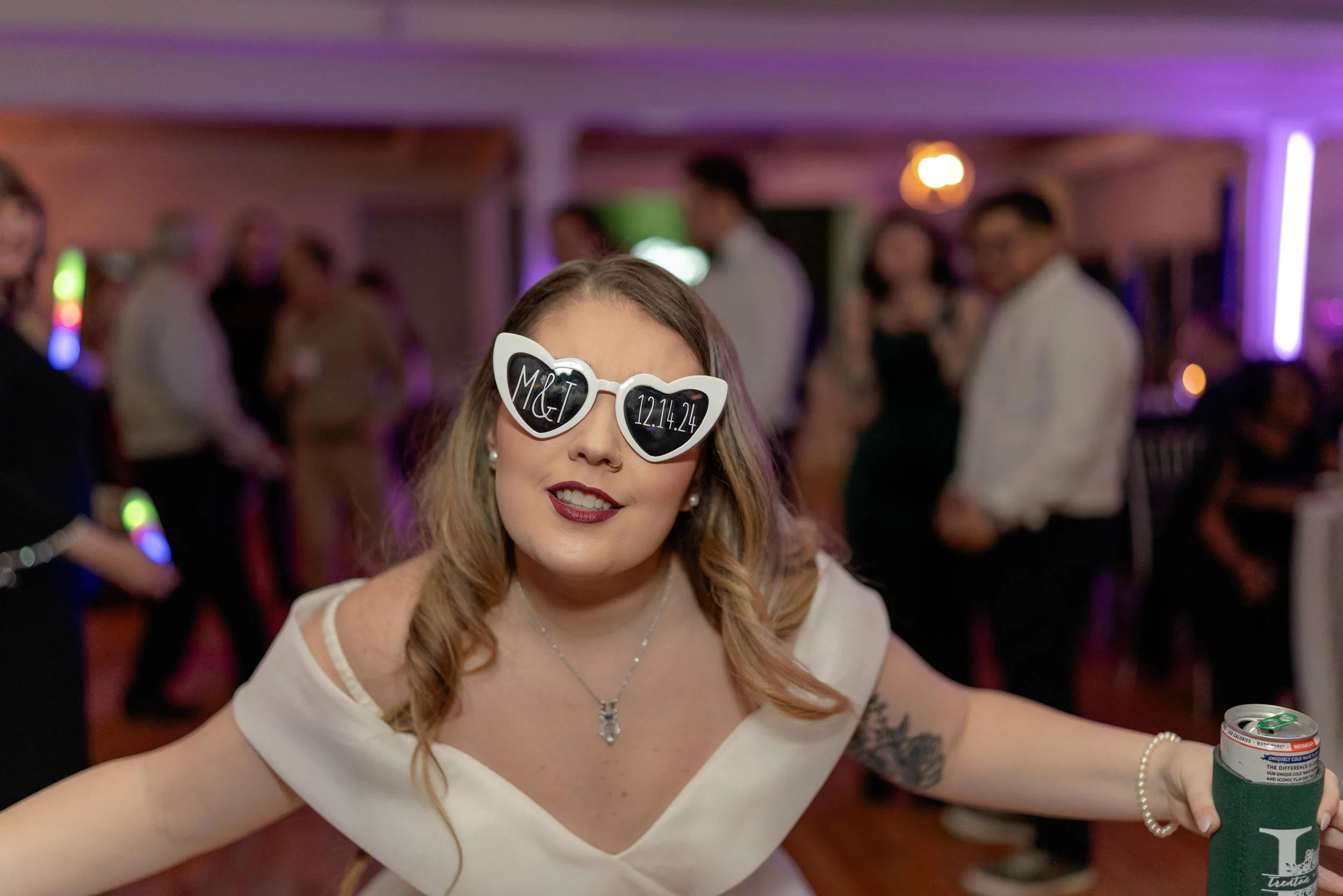 Woman in heart-shaped sunglasses at a party, wearing a white off-shoulder dress, holding a beverage can with a koozie, with people and colorful lights in the background.