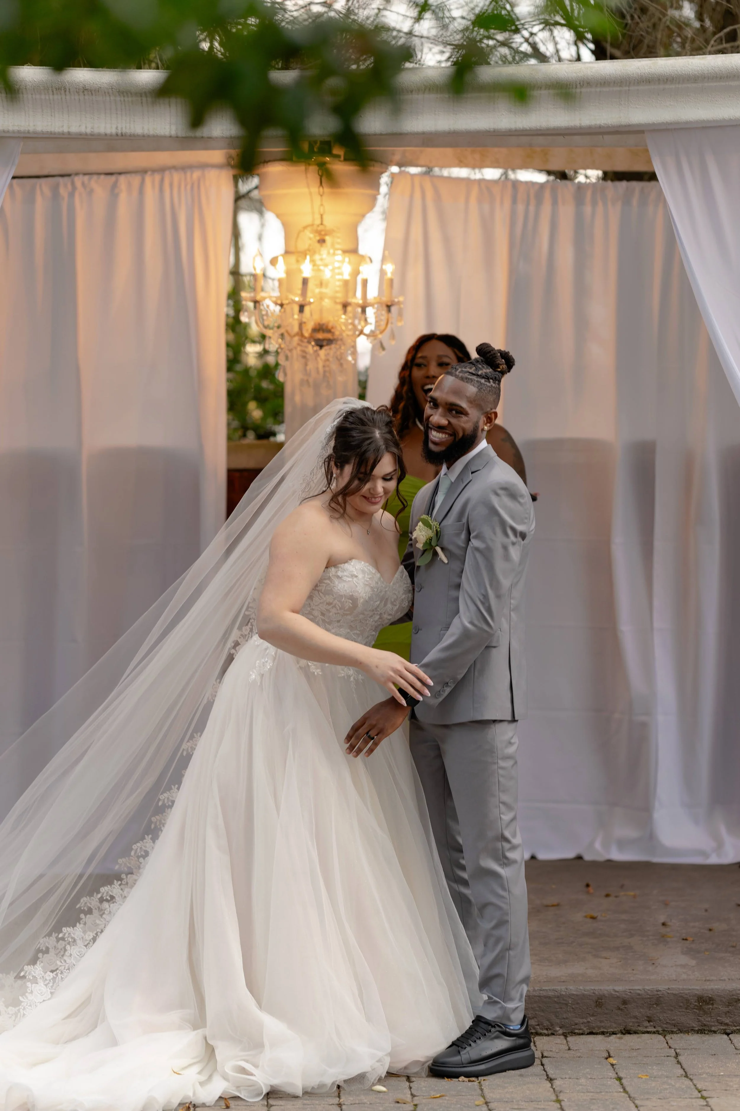 Bride and groom smiling at their wedding ceremony, with an officiant in the background under a chandelier.