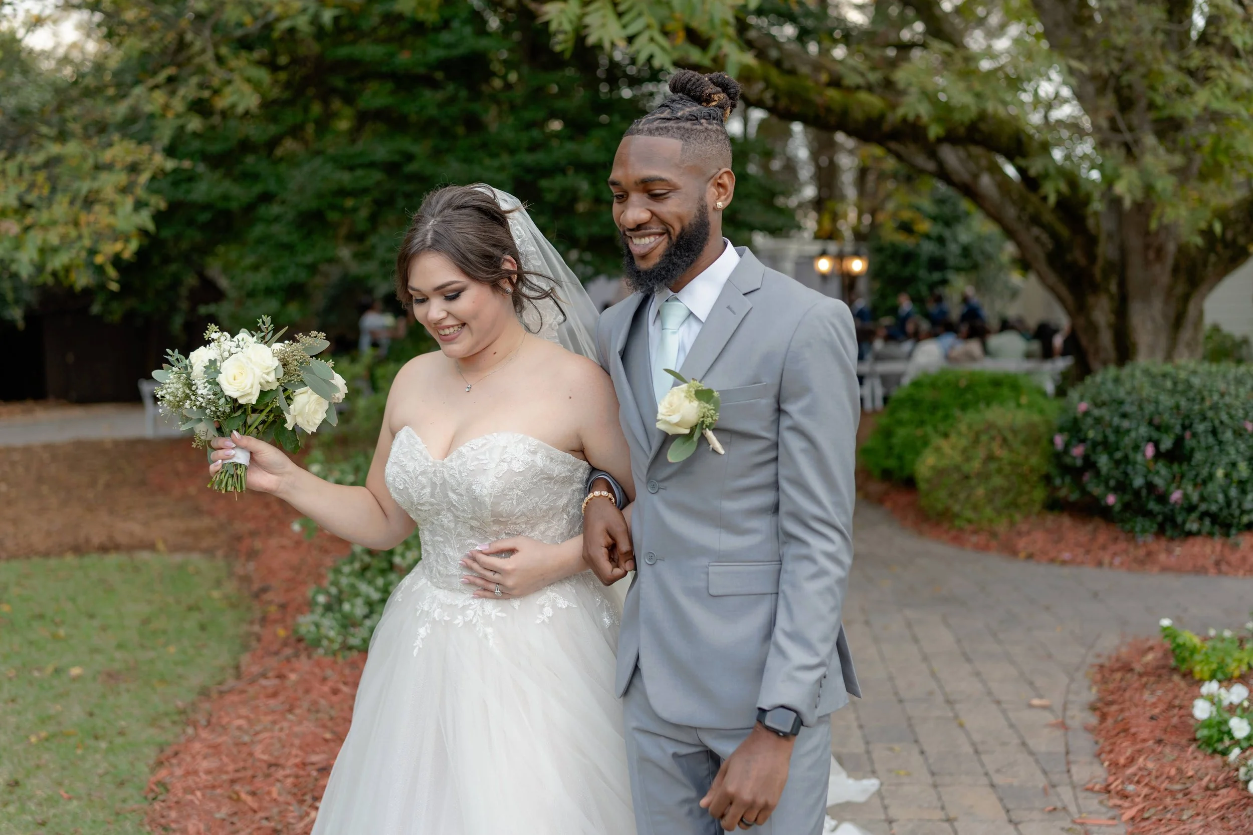 A bride and groom walking outdoors, the bride wearing a white wedding dress and holding a bouquet, the groom in a light gray suit, both smiling.