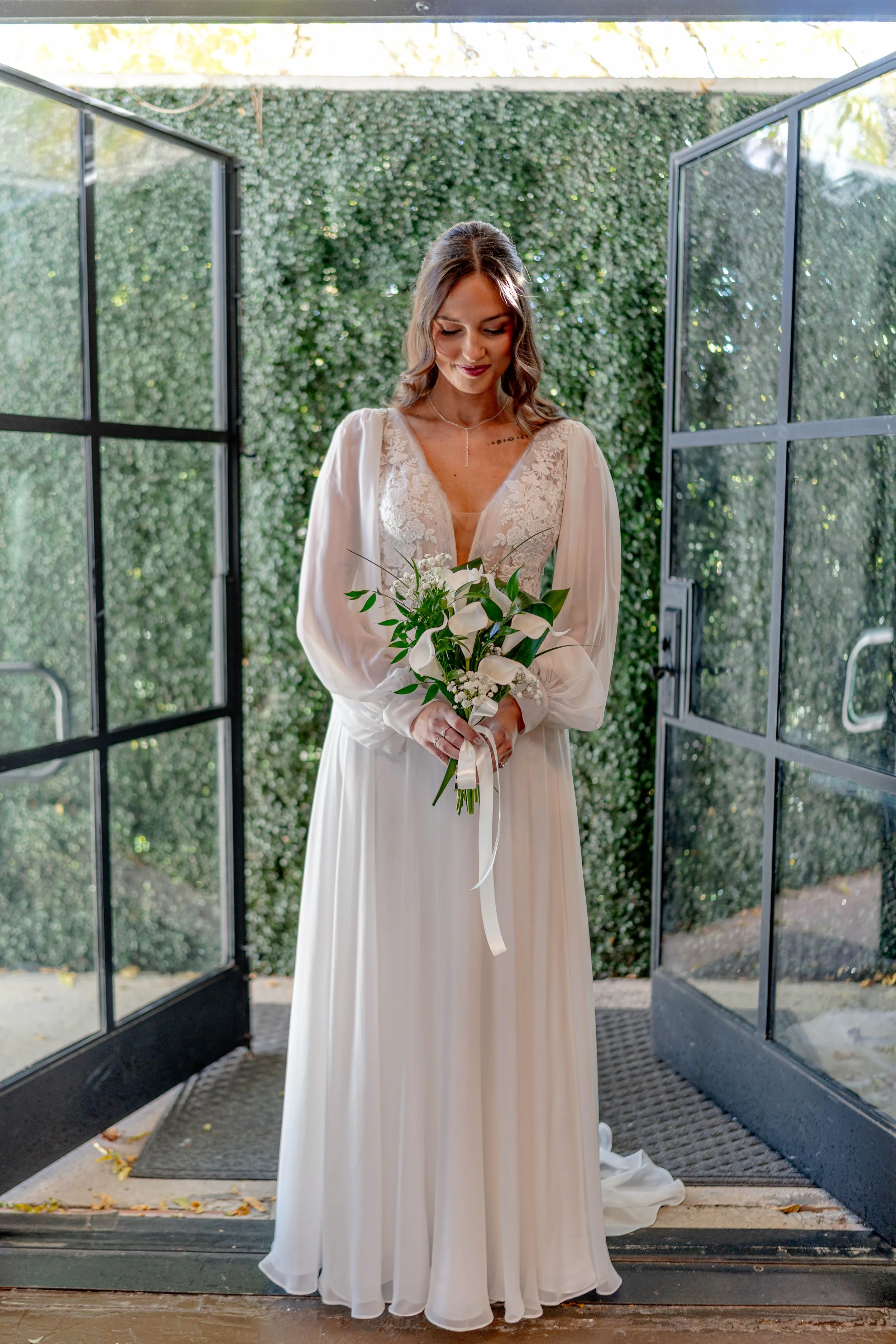 A woman in a white wedding dress holding a bouquet of white calla lilies and greenery, standing in doorway with open glass doors and a green leafy wall behind her.