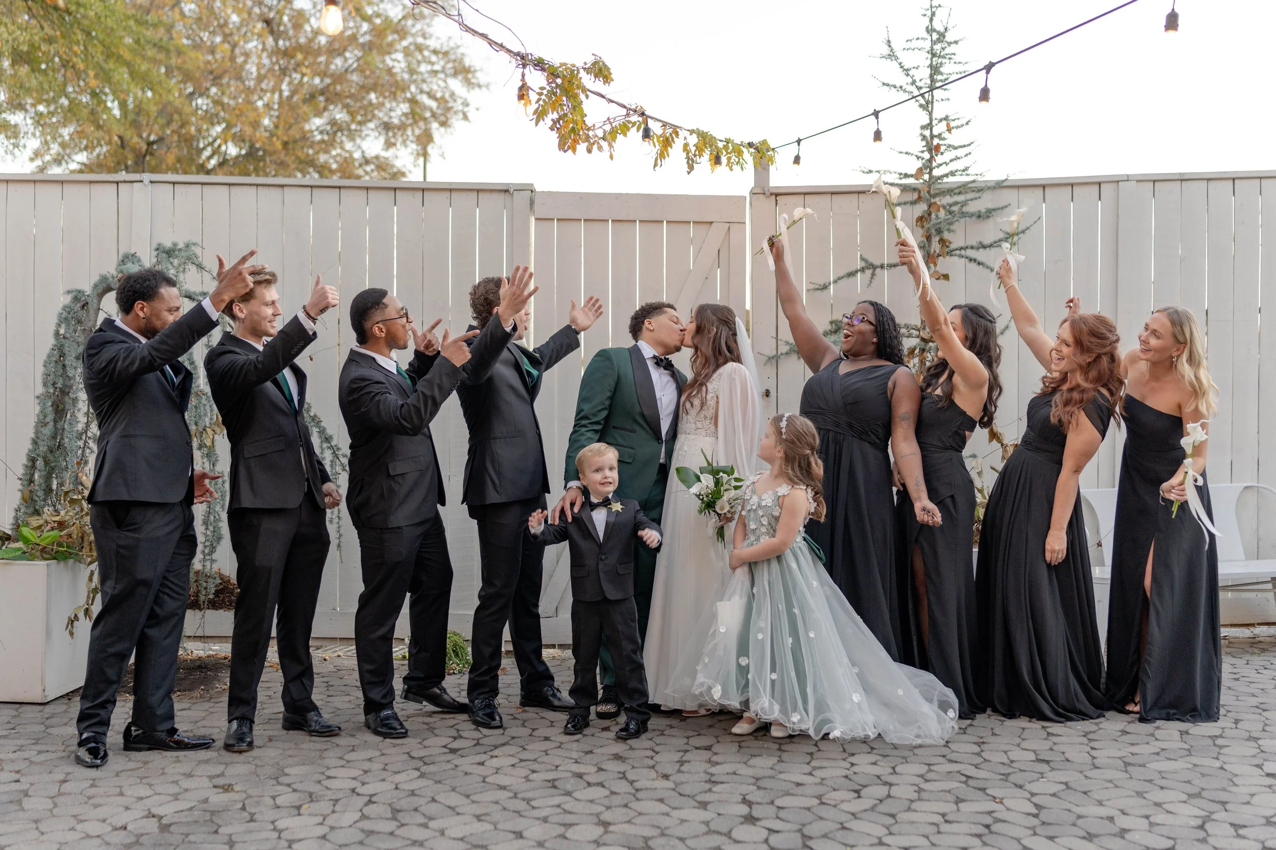 A wedding celebration with numerous guests gathered outdoors, watching and cheering the couple as they kiss. The bride and groom are at the center, surrounded by friends and family, with some holding white calla lilies.