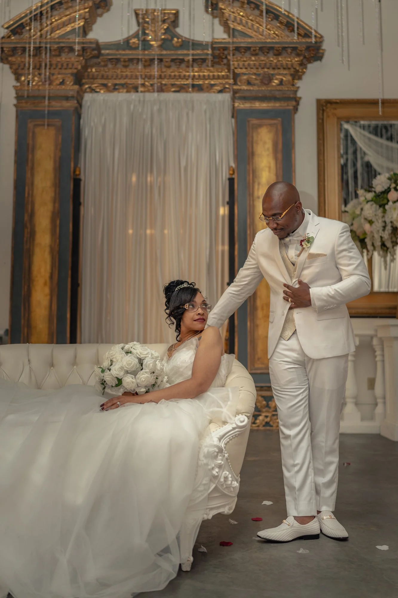 Bride and groom in white attire, bride holding bouquet, elegant setting with ornate backdrop.