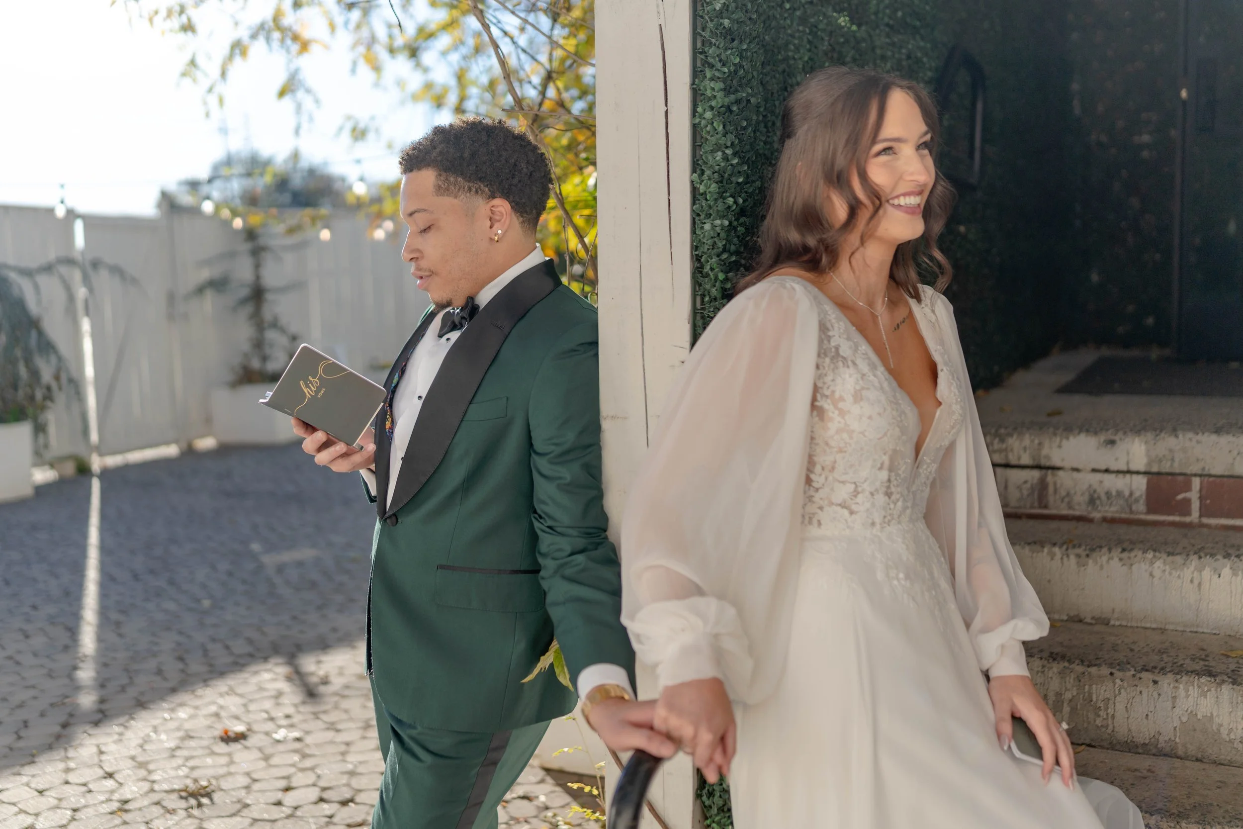 A bride and groom standing apart, holding hands. The groom is looking at a small book, and the bride is smiling, leaning against a wall. They are dressed in wedding attire outdoors during daytime.