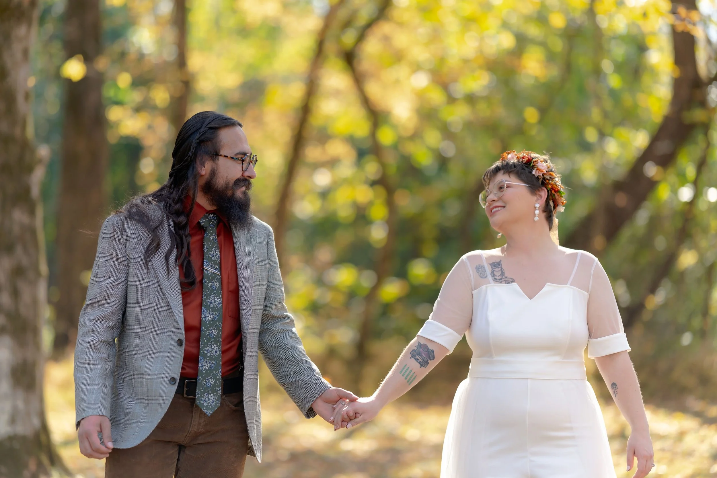 A couple holding hands outdoors, with the man in a suit and the woman in a white dress, surrounded by trees and sunlight.