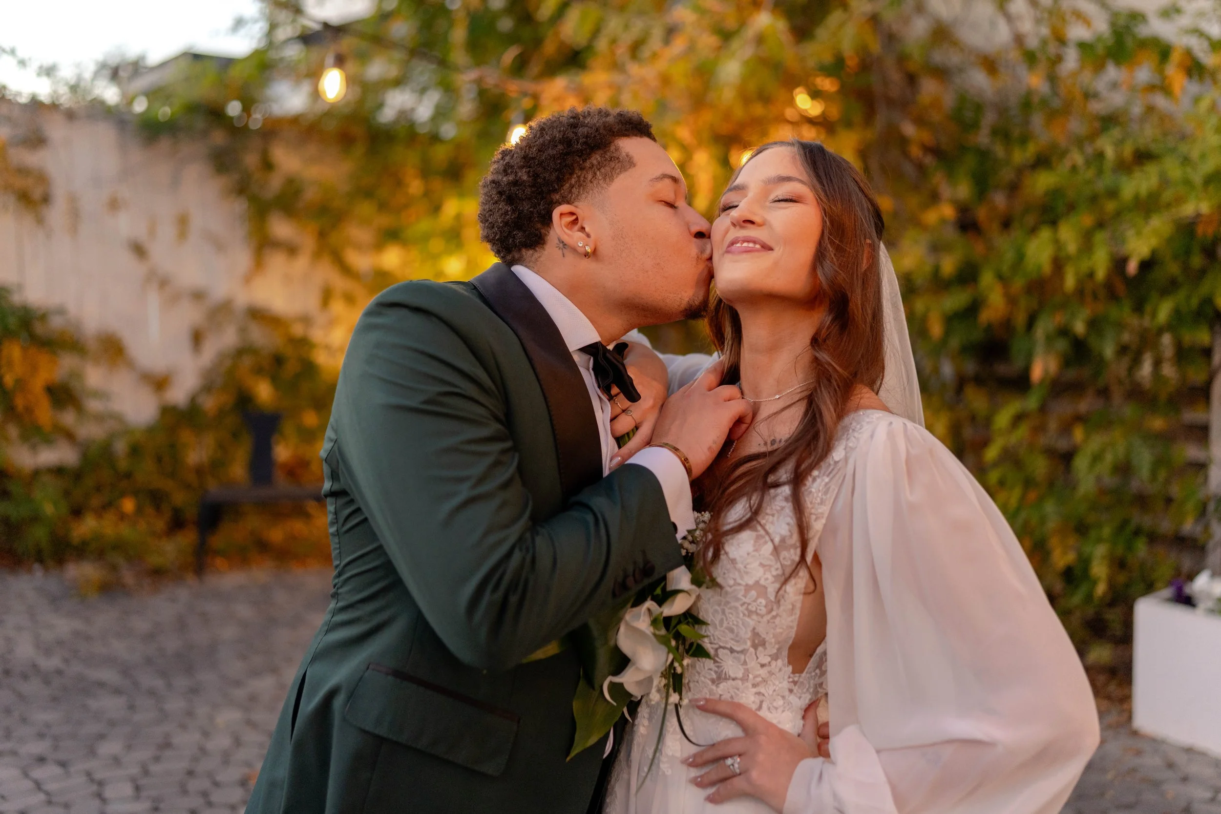 A bride and groom sharing a kiss outdoors during fall, with trees and string lights in the background.