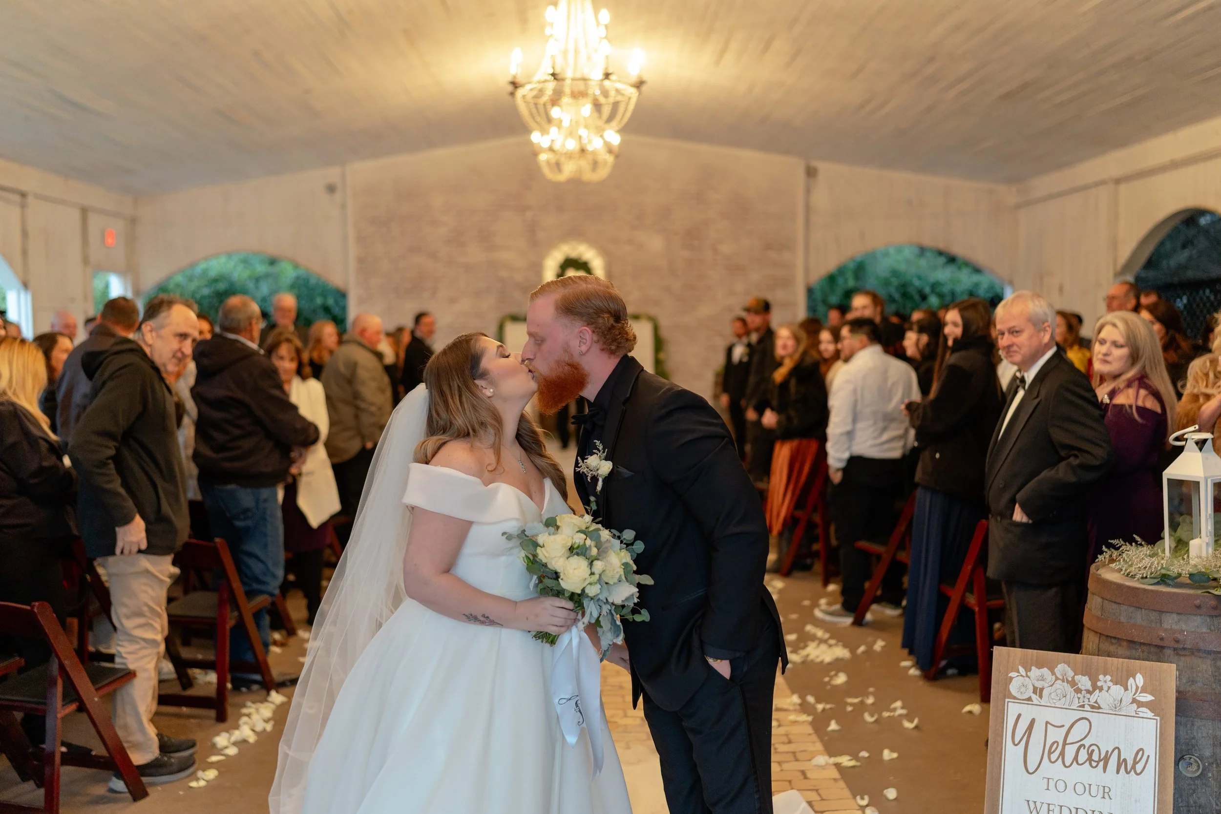 Bride and groom kissing in a wedding ceremony venue, surrounded by guests, with a "Welcome to our wedding" sign nearby.