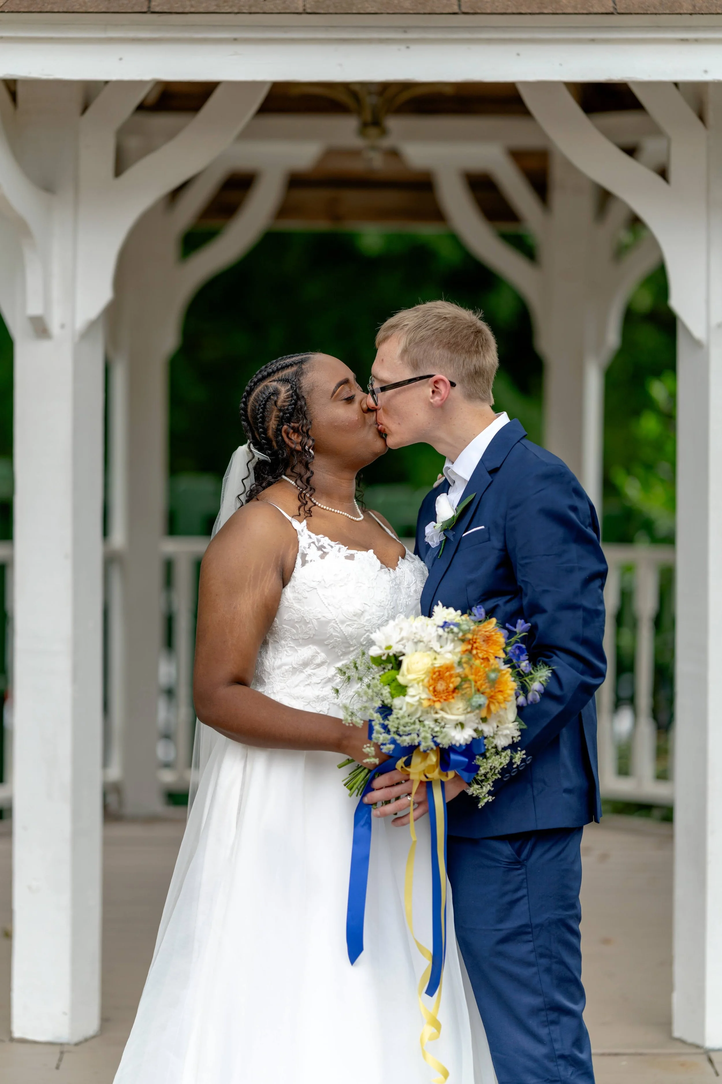 A bride and groom kissing under a gazebo. The bride is wearing a white dress and holding a colorful bouquet, while the groom is in a blue suit.