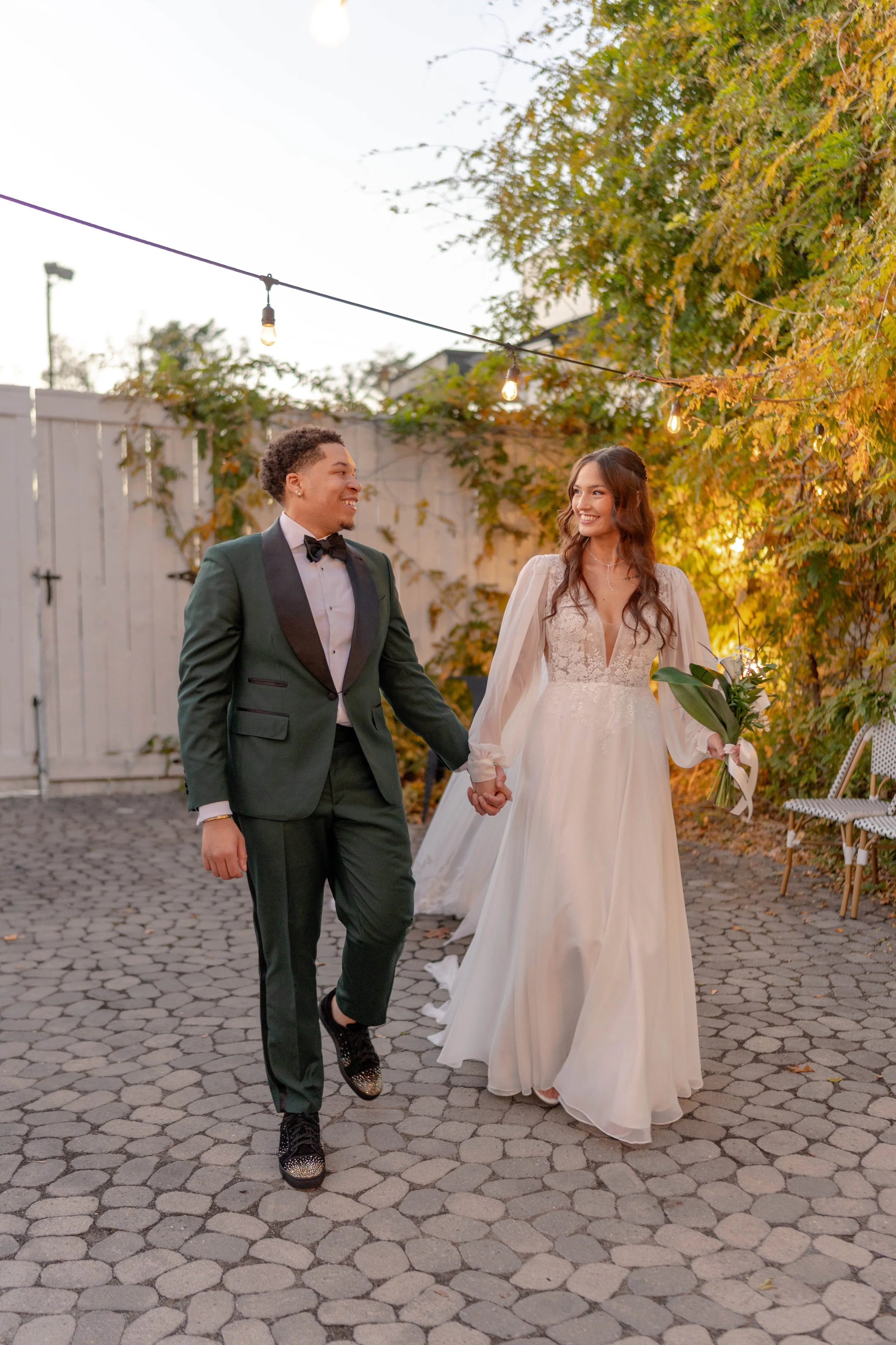 A couple, dressed for a wedding, walking hand-in-hand outdoors during evening, with autumn trees and string lights overhead, smiling at each other.