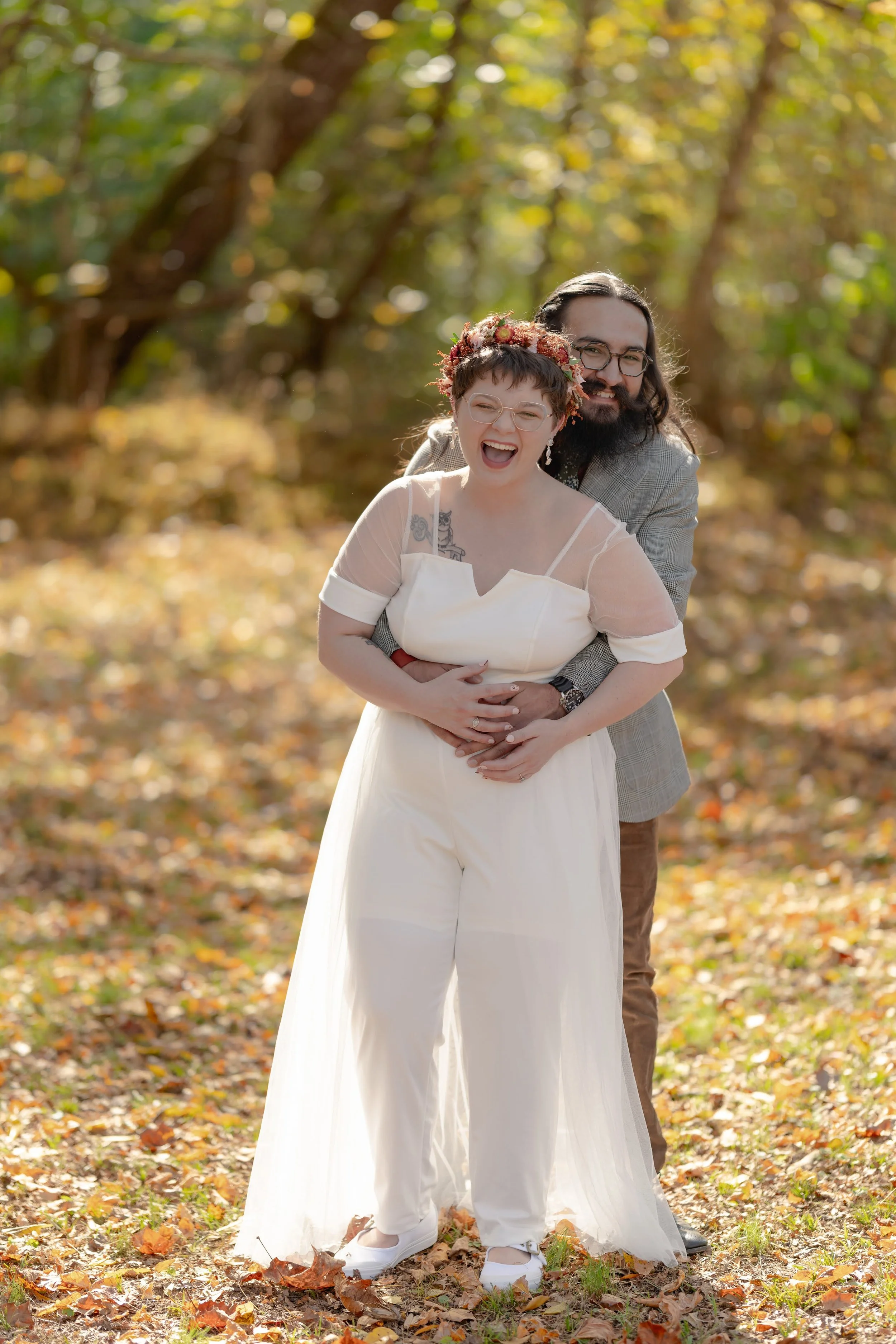 A couple laughing and embracing in a sunny forest, with the woman wearing a white dress and floral crown and the man wearing glasses and a suit.