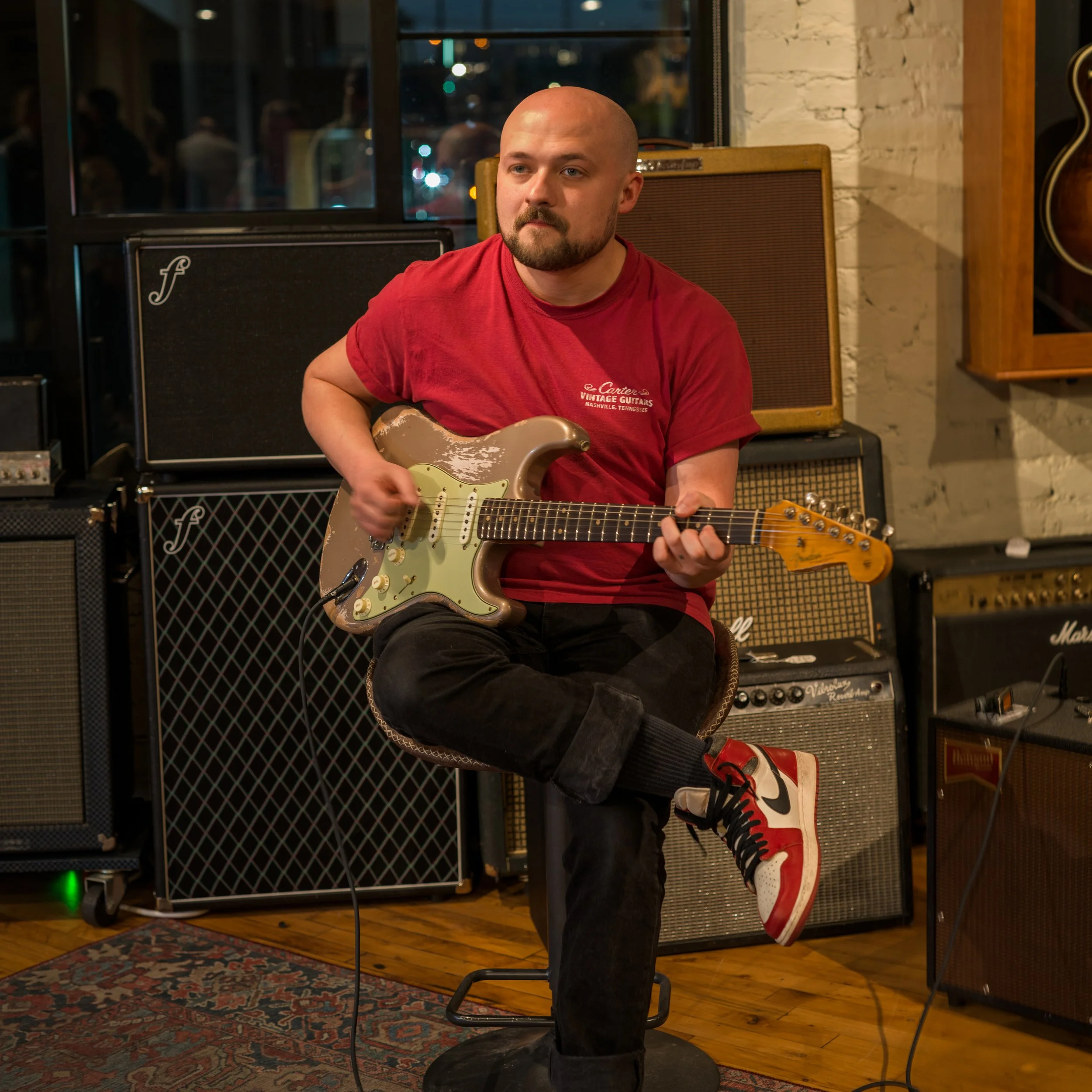 man playing a beat-up Fender guitar, sitting in front of several amps
