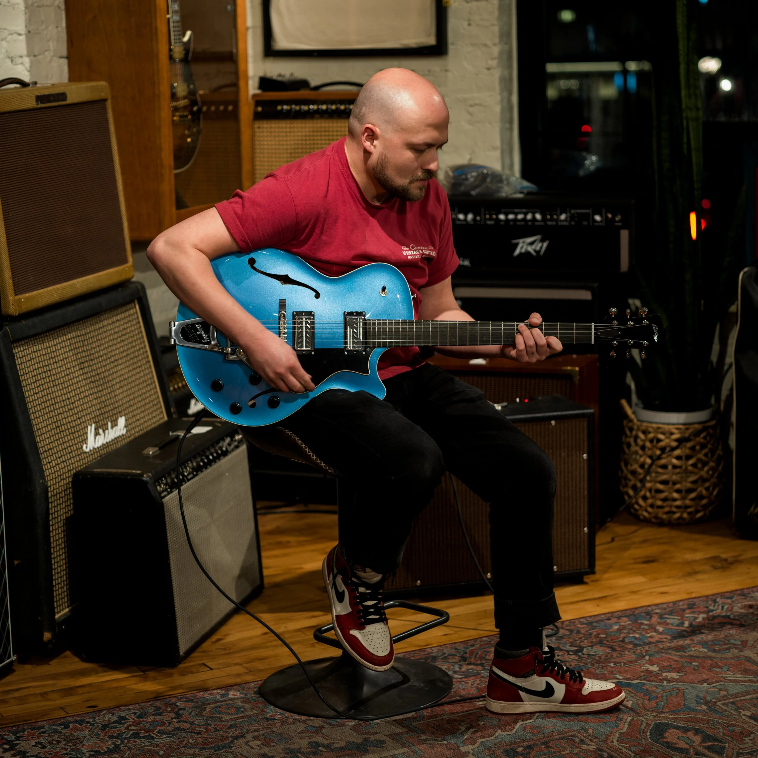 Man playing blue Collings guitar, seated in front of an assortment of amps.