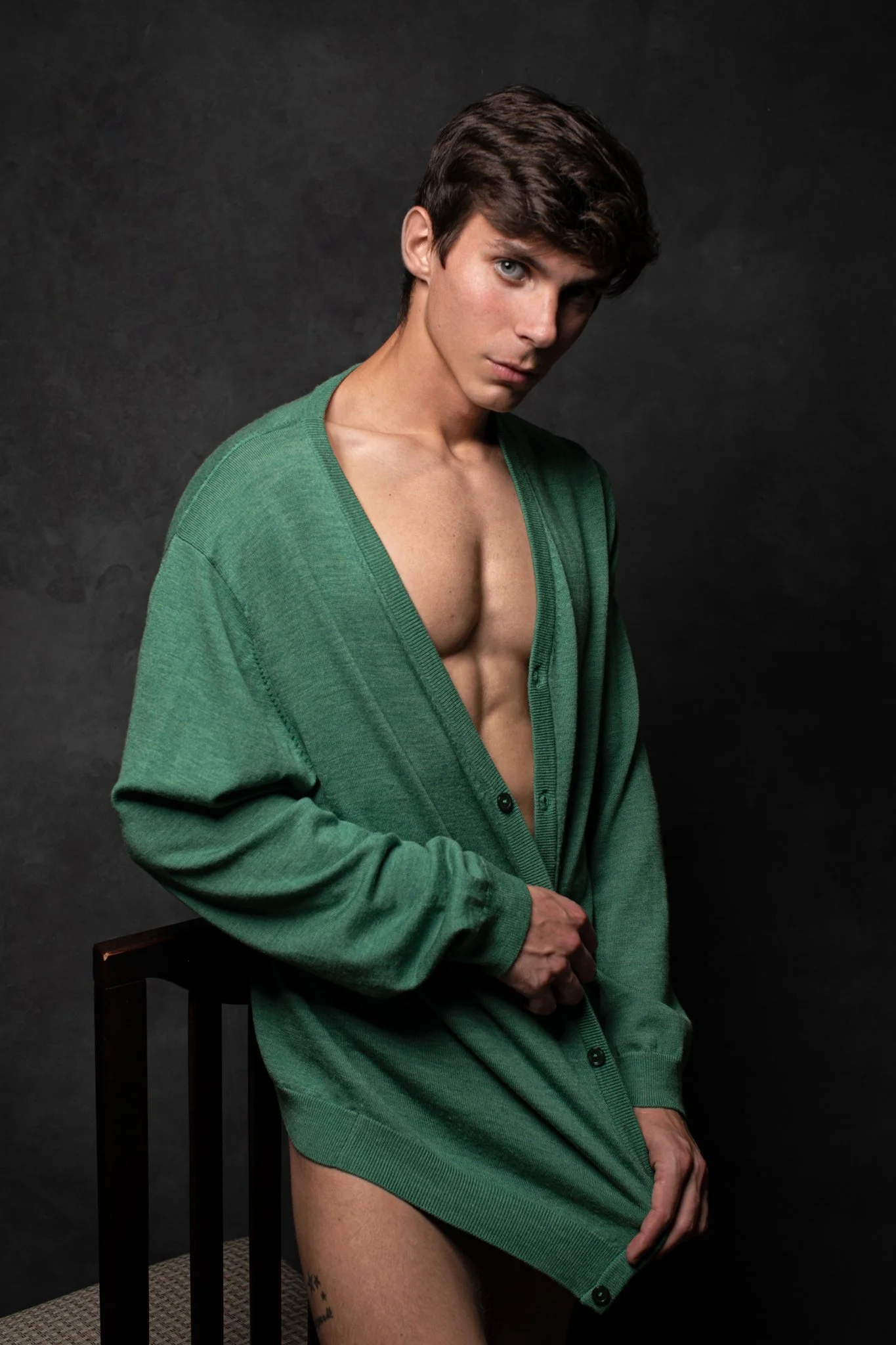 A young man with brown hair and blue eyes wearing an oversized green cardigan, partially open, revealing his bare chest and abdomen, sitting on a wooden chair against a dark background.