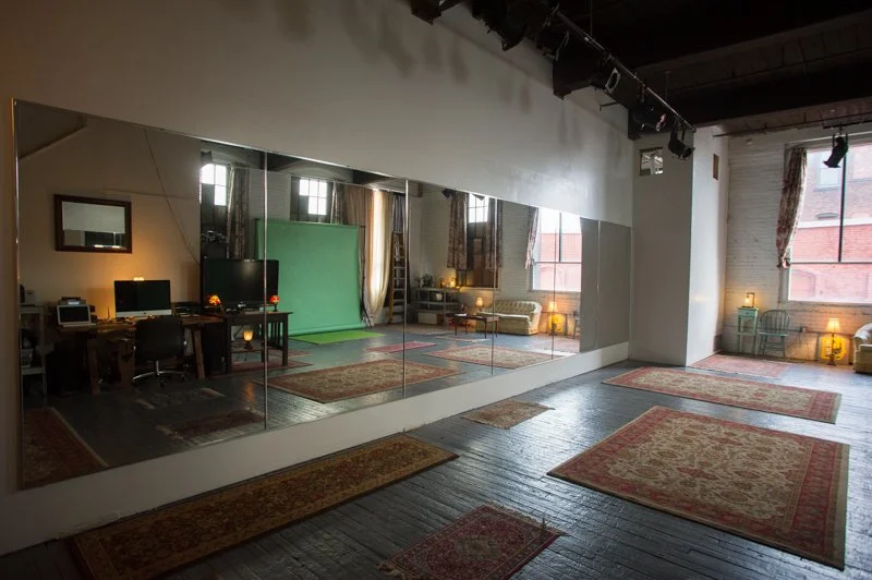 A spacious room with a large mirror wall, hardwood floors covered with multiple oriental rugs, large windows with curtains, and a mix of furniture including desks, chairs, a sofa, and a green screen backdrop.