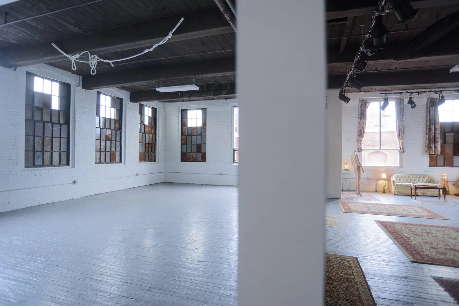 Split view of two interiors of large, industrial-style lofts with large windows, white painted brick walls, and wooden floors. The left side shows an empty room with graffiti-covered windows, dark ceiling beams, and a loose hanging rope. The right si