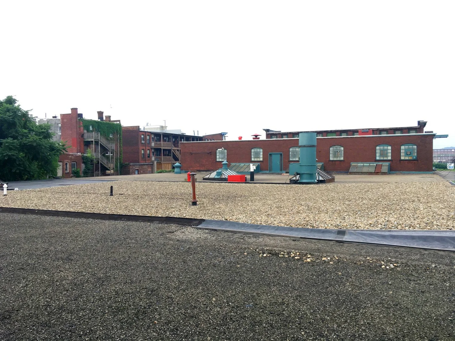 Rooftop view of brick buildings with HVAC systems and a gravel surface, some windows with green trim, and a tree on the left side under an overcast sky.