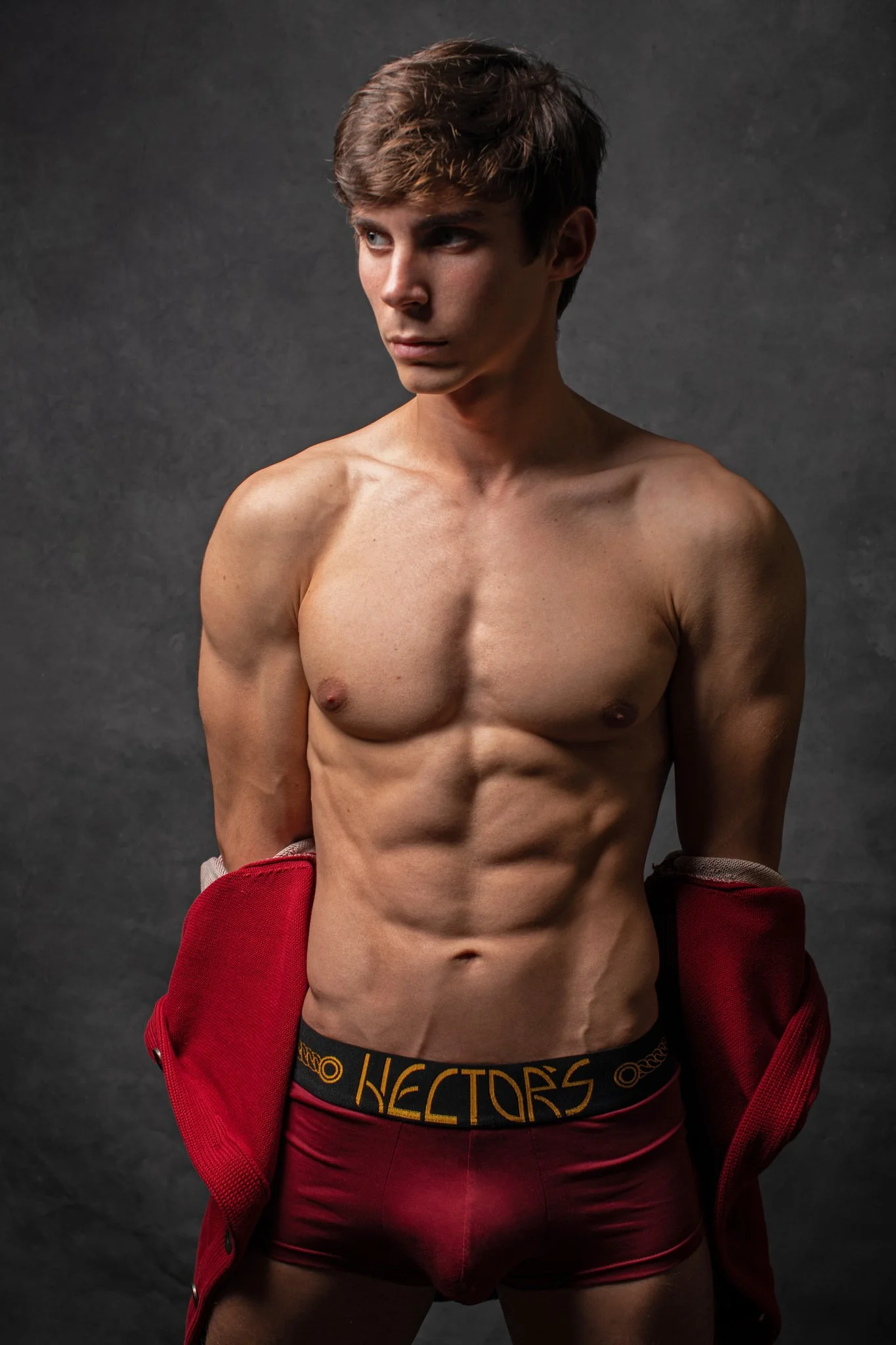 A shirtless young man with a muscular build, wearing red underwear and a partially open red jacket, standing against a dark background.